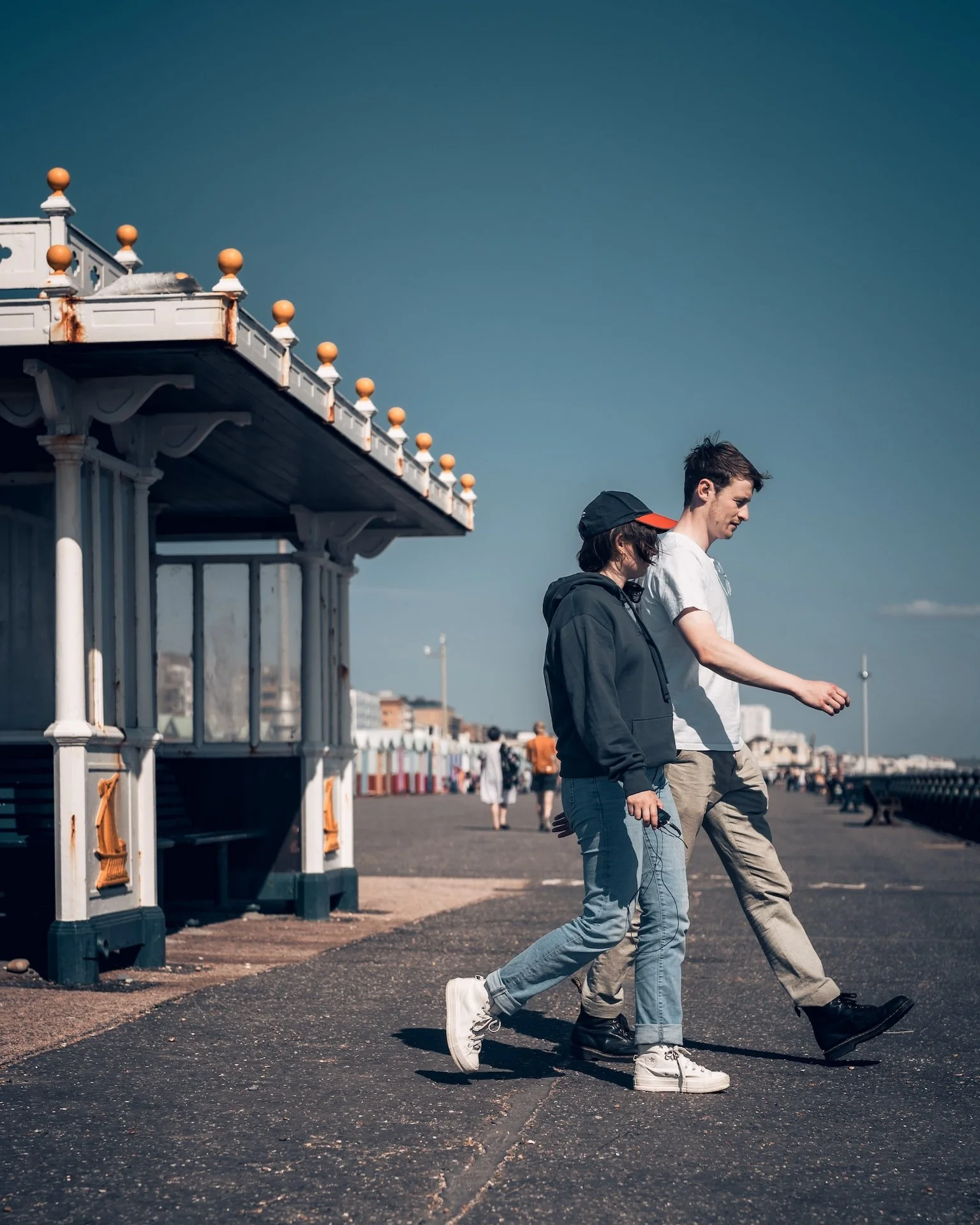 Two young men walk along a boardwalk near the beach, with a pier extending into the ocean on the left side of the image. The sky is clear and blue, and there are other people in the background.