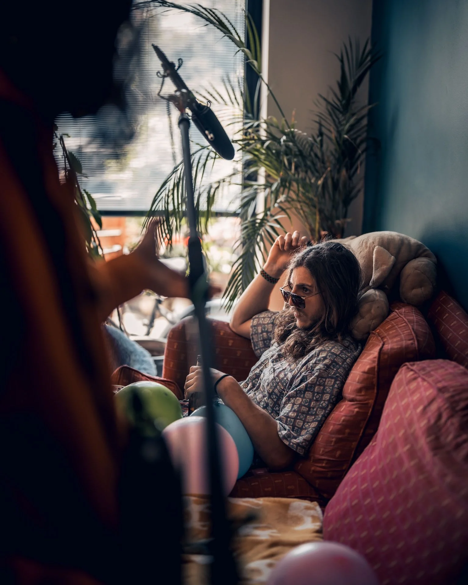 A woman with long hair and glasses relaxing on a patterned couch surrounded by pillows, with a large plant and window with blinds behind her, as someone adjusts a microphone stand nearby.