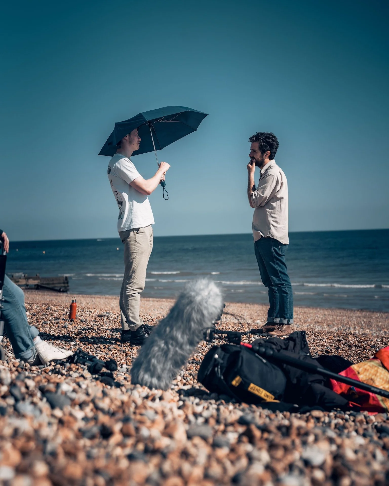 Two men standing on a pebble beach, one holding an umbrella over the other, with the ocean in the background, and a dog sitting in the foreground.