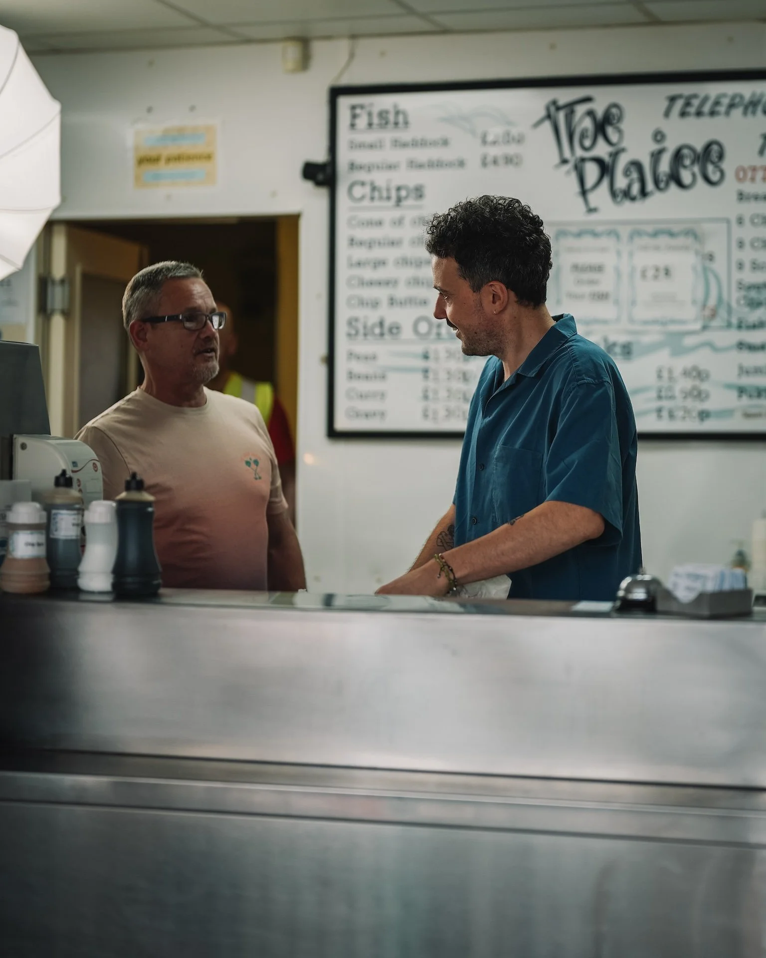 Two men are having a conversation inside a fish and chips shop, with a menu board listing various fish and chips options in the background.