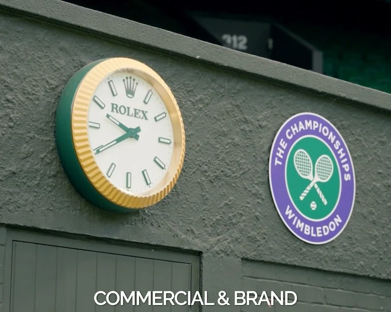 A wall-mounted Rolex clock showing the time as 3:50, next to a sign for 'The Championships Wimbledon' tennis tournament, on a gray textured wall.