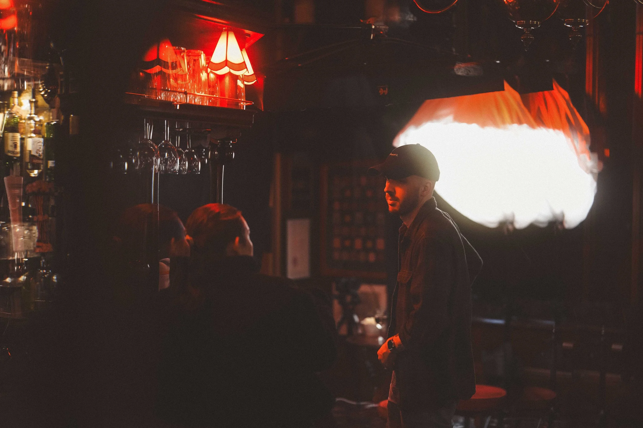 A dimly lit bar with red lighting, wine glasses hanging upside down, and a man wearing a cap and jacket standing near the bar, engaged in conversation or observation.