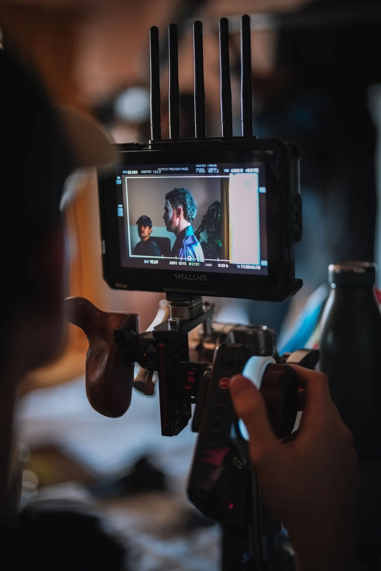 A camera operator films two men sitting indoors, one with gray hair and the other with dark hair, through a professional camera monitor.