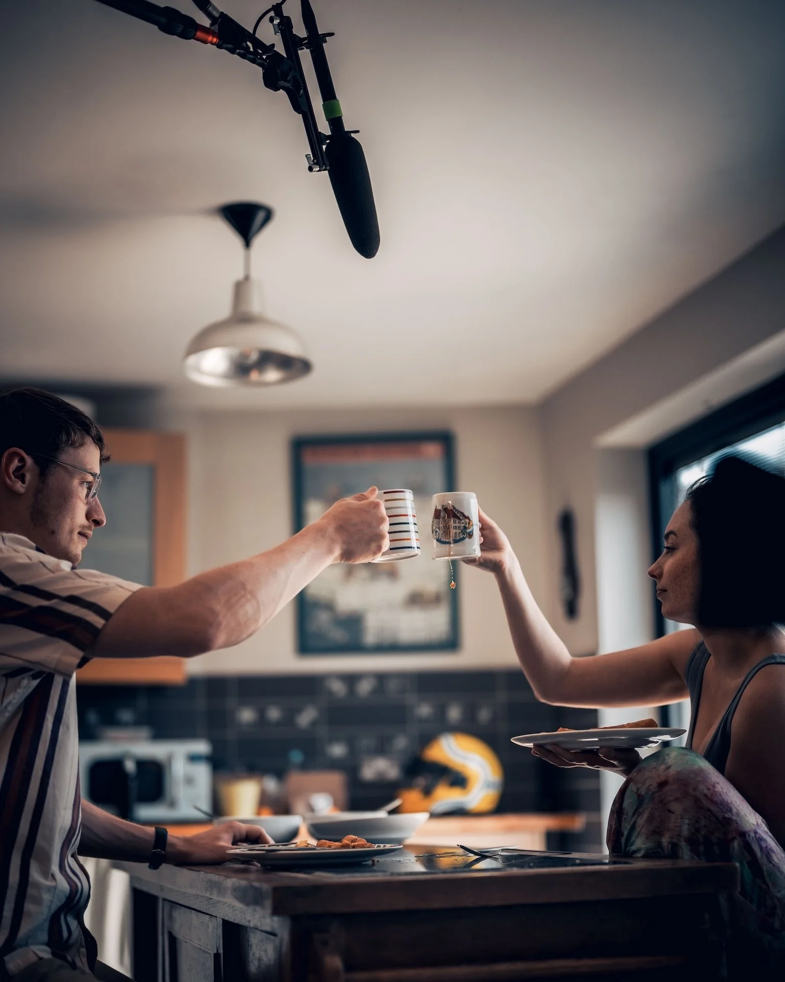 Two people raising coffee mugs in a toast in a kitchen with a man sitting at the counter with a plate of food and a woman holding a plate, large window, ceiling light, and a framed picture in the background.