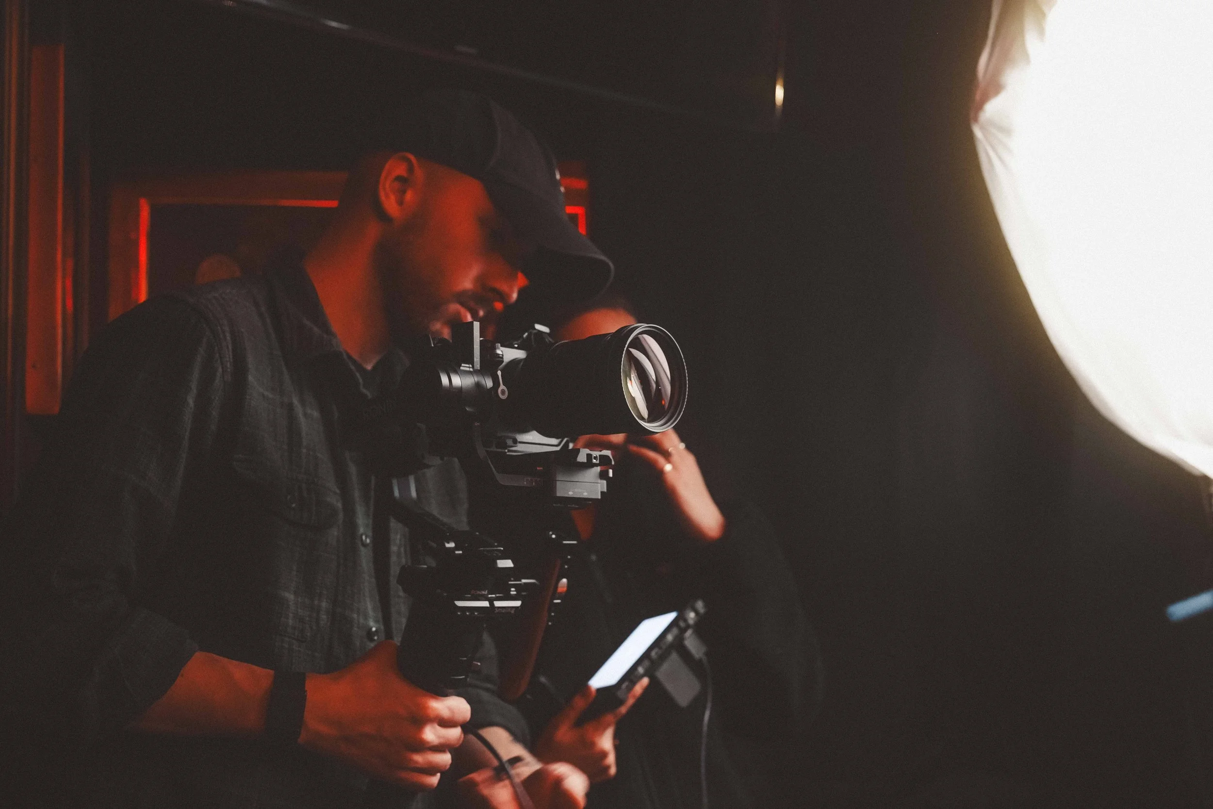 A person wearing a dark cap, black shirt, and plaid jacket is operating a camera on a tripod in a dimly lit environment with red lighting, with another individual partially visible in the background.