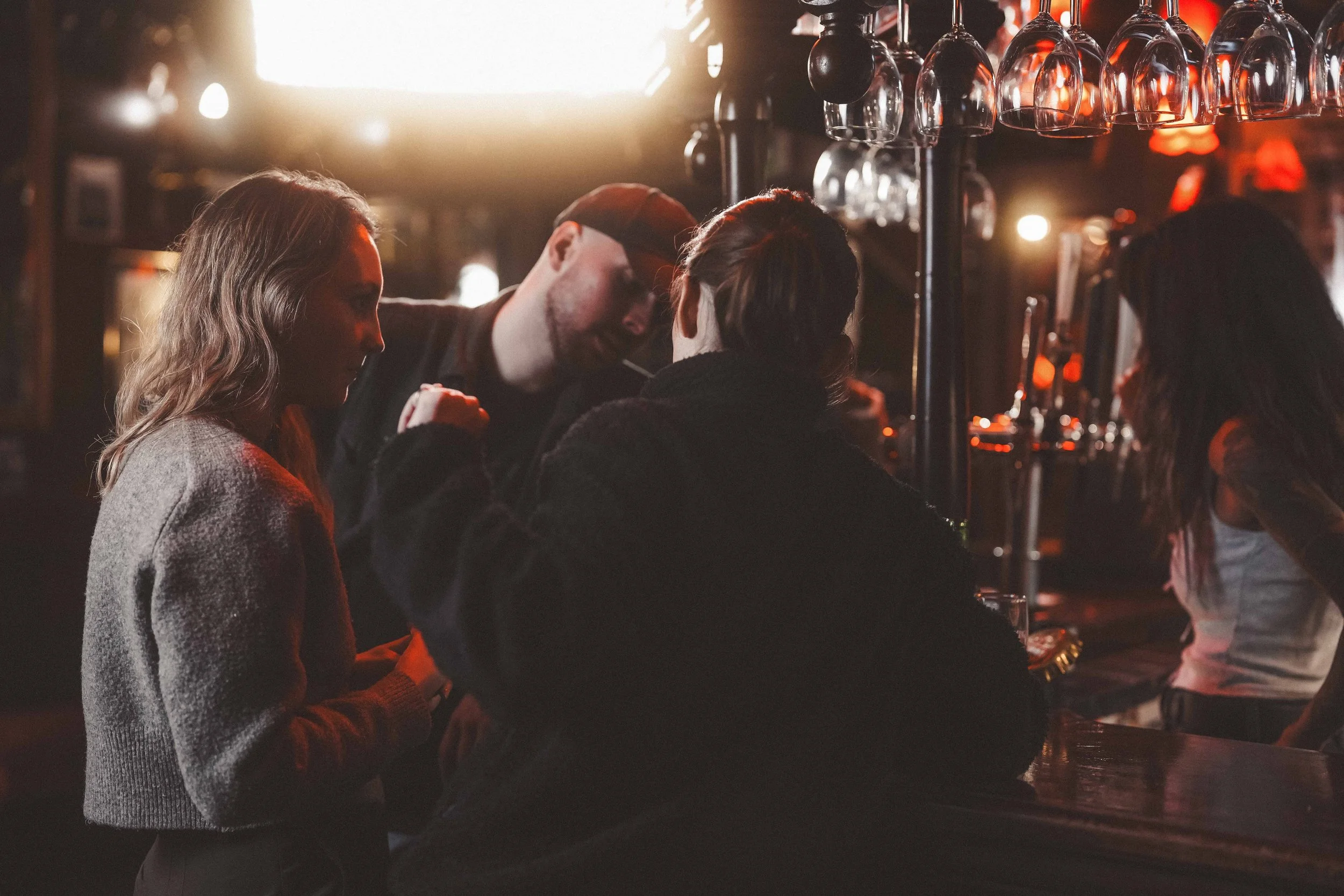 Group of people socializing at a bar with glasses hanging overhead in a dimly lit setting.