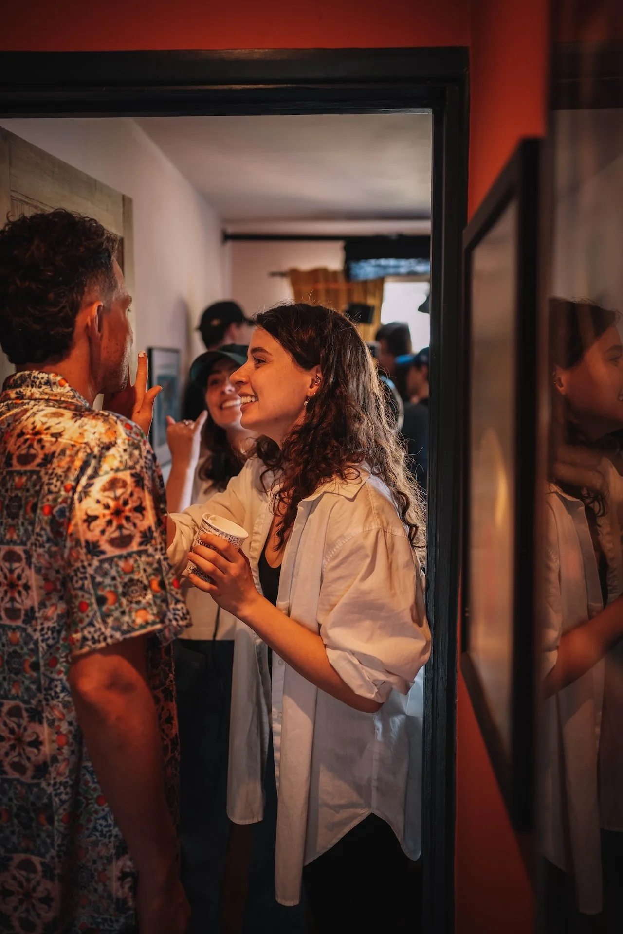 A group of young adults socializing indoors, with a woman holding a yogurt container and talking to a man in a patterned shirt. Others are visible in the background.