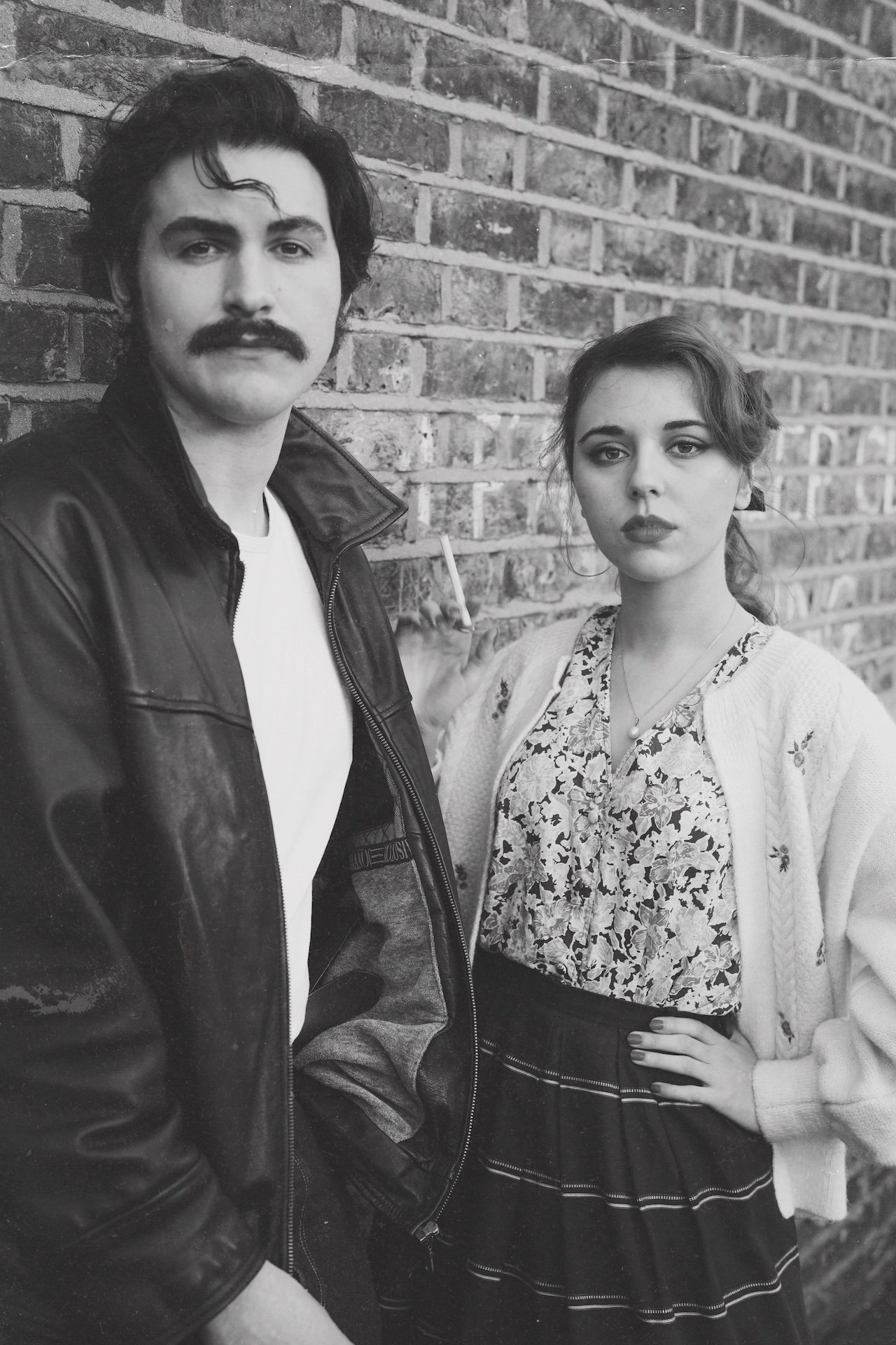 A black and white photo of a man with dark hair and a mustache, wearing a leather jacket, and a woman with wavy hair, wearing a patterned blouse and a light cardigan, standing against a brick wall.