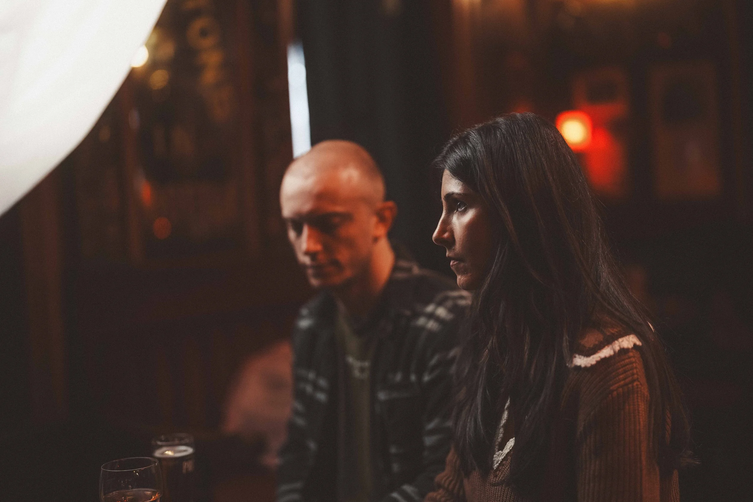 A woman with long dark hair and a man with a shaved head sitting at a dimly lit bar or restaurant, with glasses of wine in front of them, and blurred warm lighting in the background.