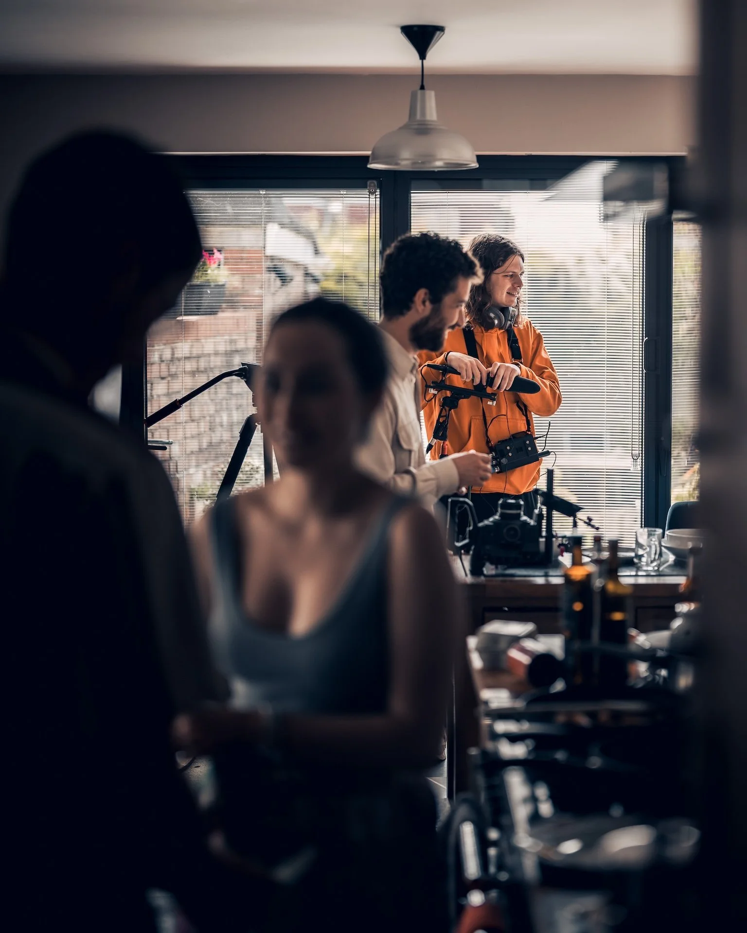 Several people in a room around a table with equipment, with one woman in an orange jacket holding a microphone and others engaging in conversation.