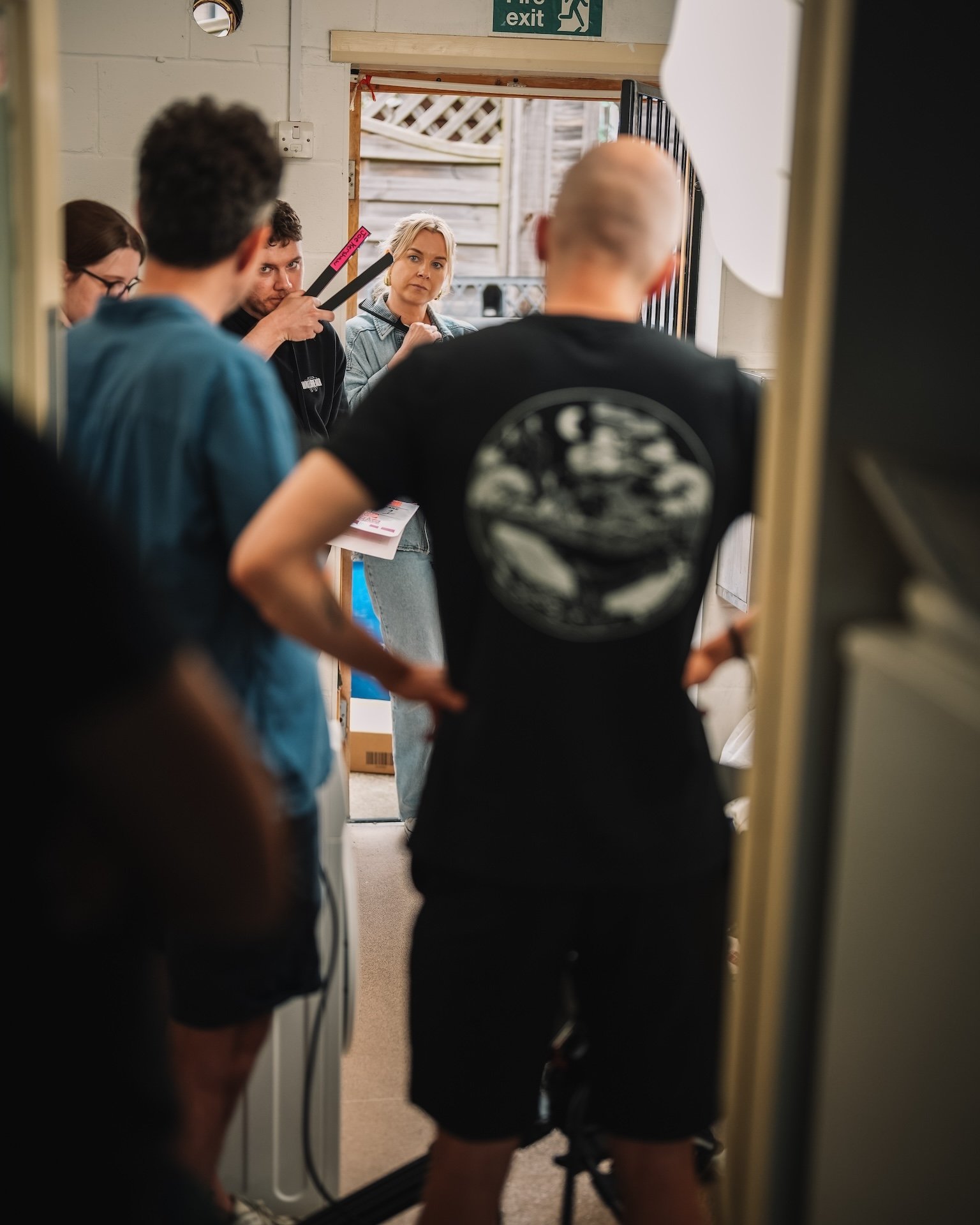 A group of young adults in casual clothing gathering near an open door, some holding skateboards, in what appears to be an indoor setting with a white brick wall and an exit sign overhead.
