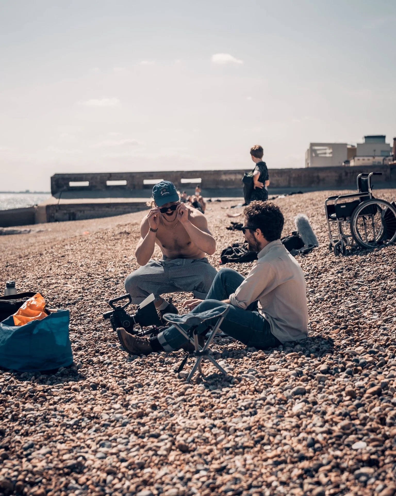 Two men sitting on a pebbled beach, one adjusting sunglasses and the other looking towards a third person standing nearby. There is a wheelchair, bags, and a small stool in the foreground, with a concrete pier and buildings in the background under a 