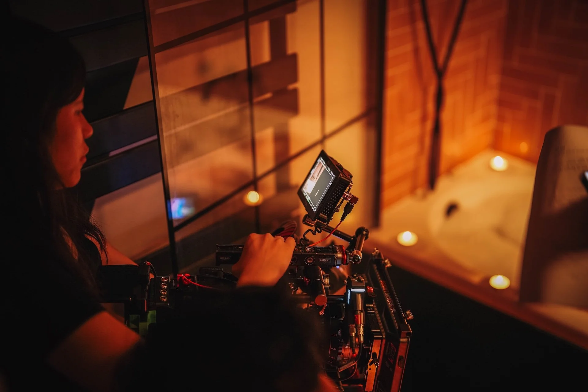 A woman operating a professional camera on a camera rig inside a warmly lit room with hanging lights and brick walls.