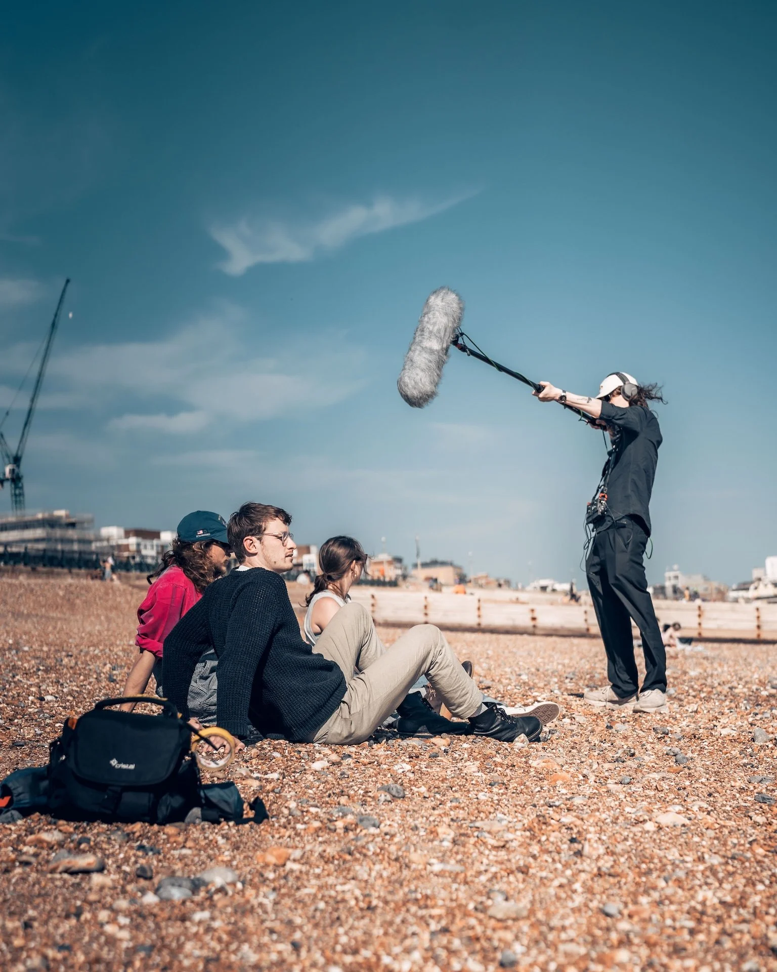 A film crew on a rocky beach filming a scene with several seated people and a person standing with a boom microphone overhead.