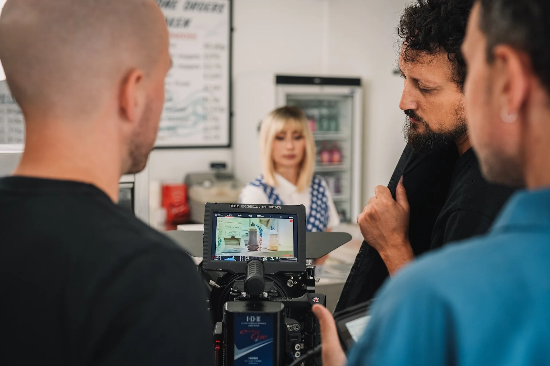 Filming of a scene using professional camera equipment in a setting with people, including a woman in the background with blonde hair and a white and blue top, and three men in the foreground, two of whom are visible from the back and one from the si
