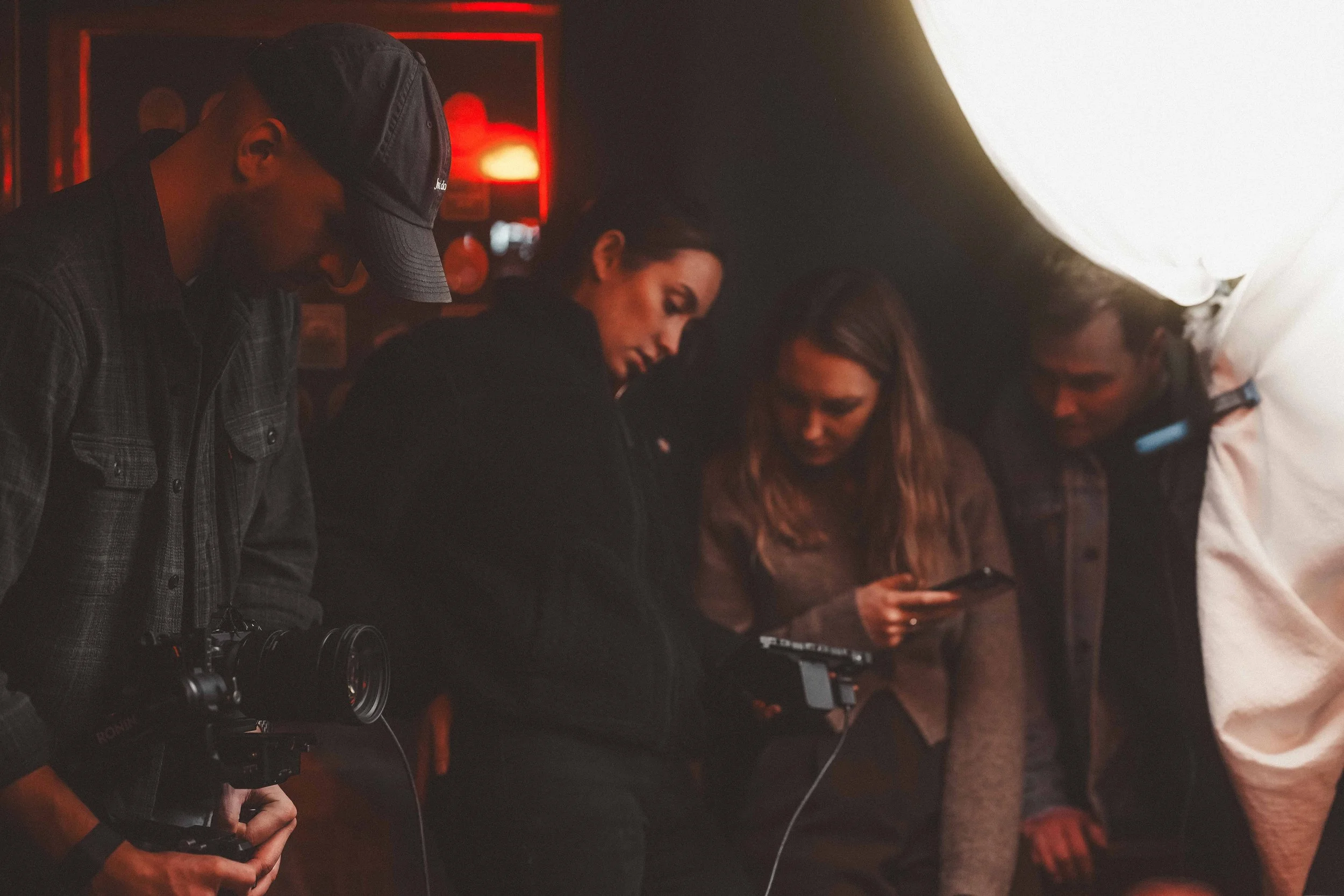 A group of four people standing closely together indoors, with some looking at a phone and others engaged, against a dimly lit background with red and warm lighting.