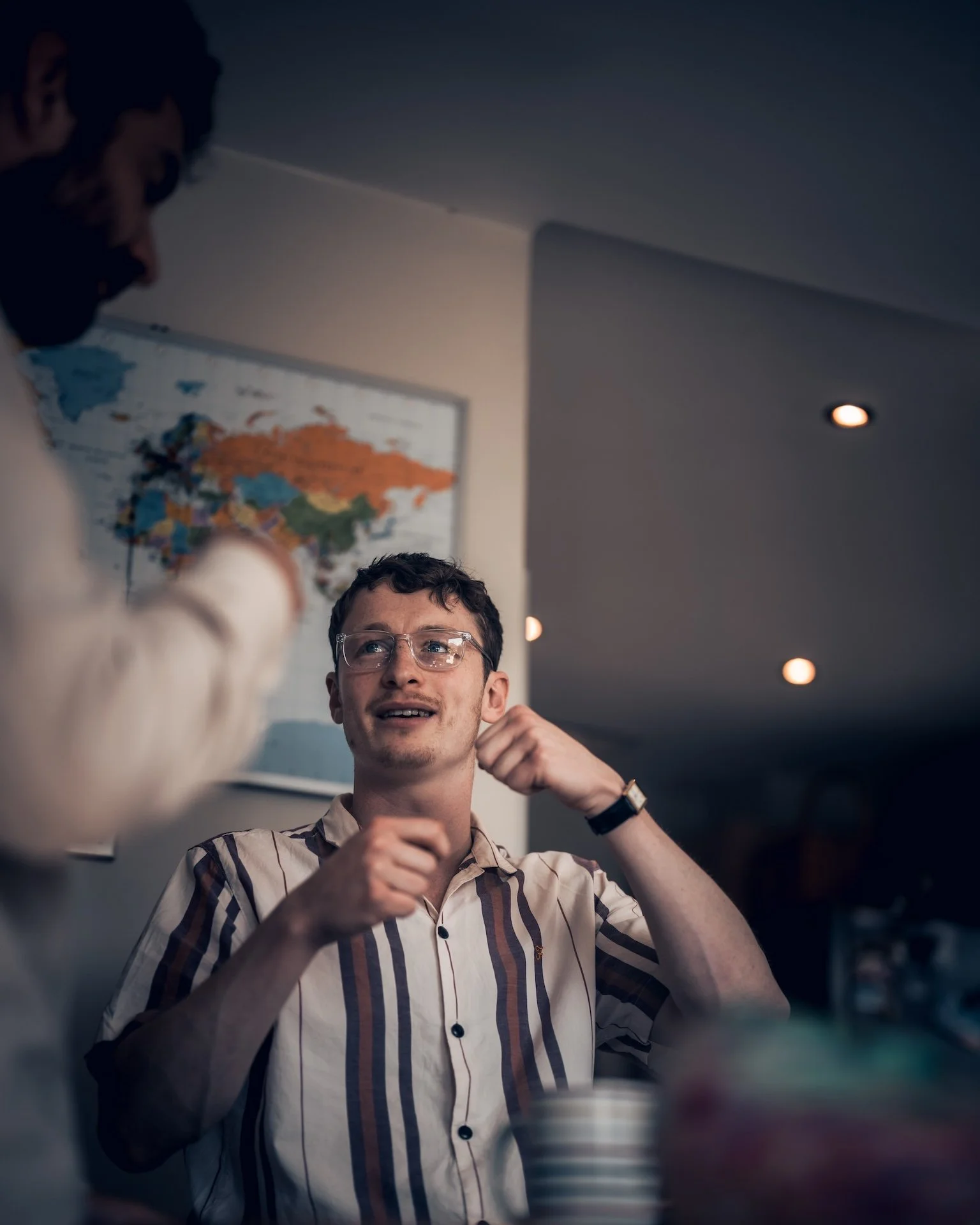 A young man with glasses and a striped shirt sitting and smiling, talking to a person whose face is not visible, in a dimly lit room with a world map on the wall.