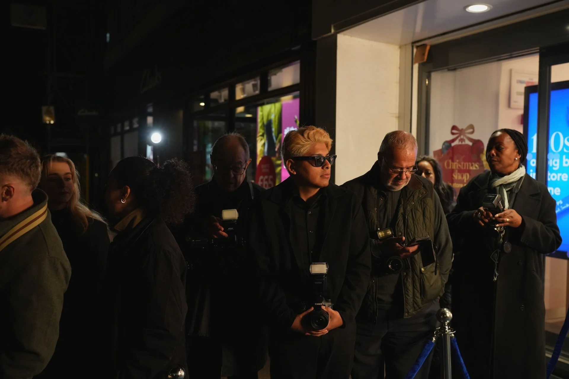 A group of people standing in a line outside a store at night, some holding cameras or phones, with a Christmas-themed sign behind them.