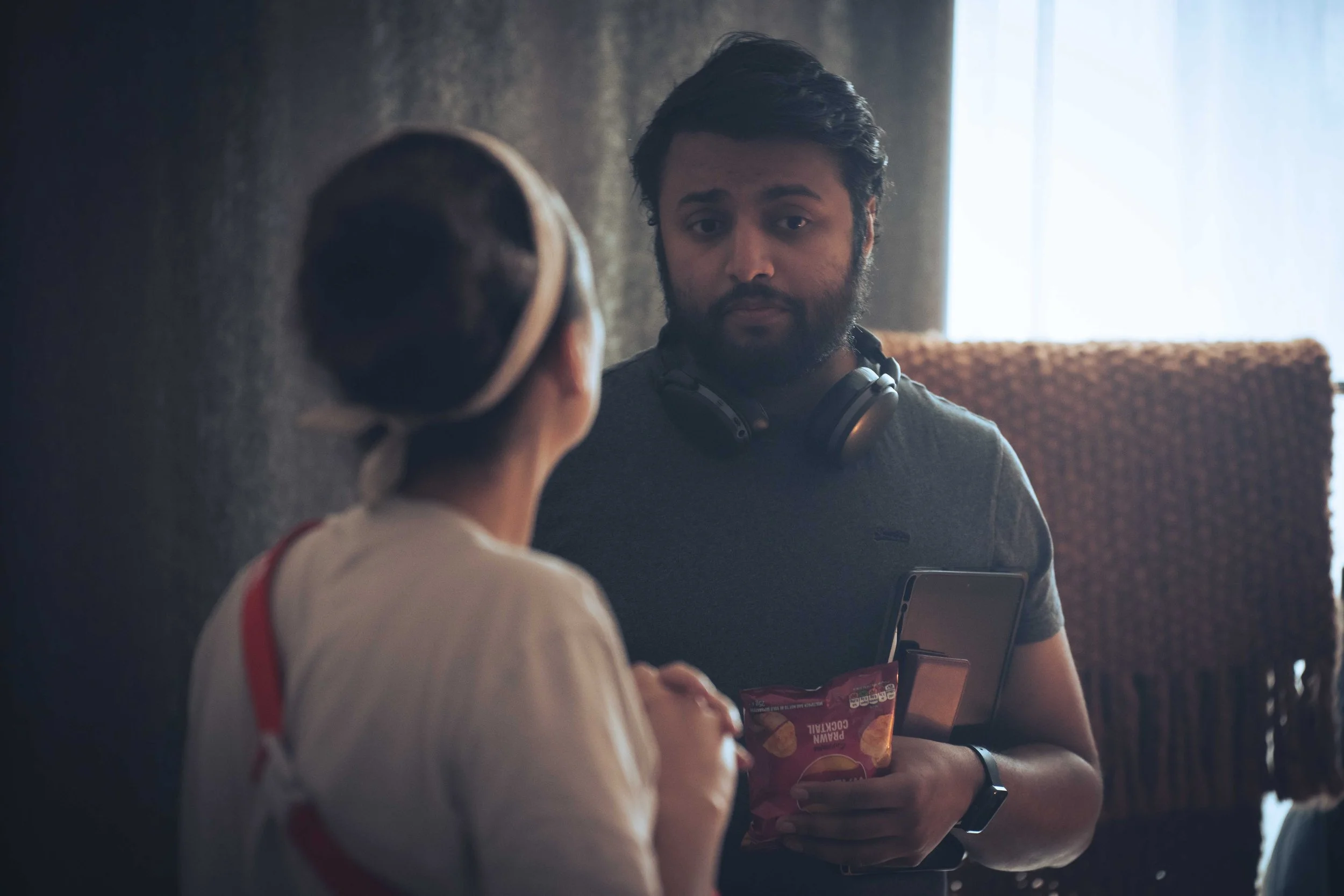 A man with a beard and dark hair wearing headphones and a watch, holding a smartphone, appears to be talking to a child. The child, seen from behind, wears a white shirt with red suspenders and has dark hair.