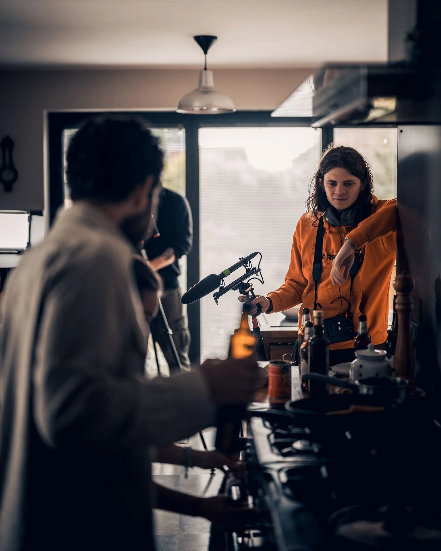 Group of people in a kitchen, one person in an orange hoodie holding a microphone boom, others sitting and talking around a table with bottles and cups, large window in the background.