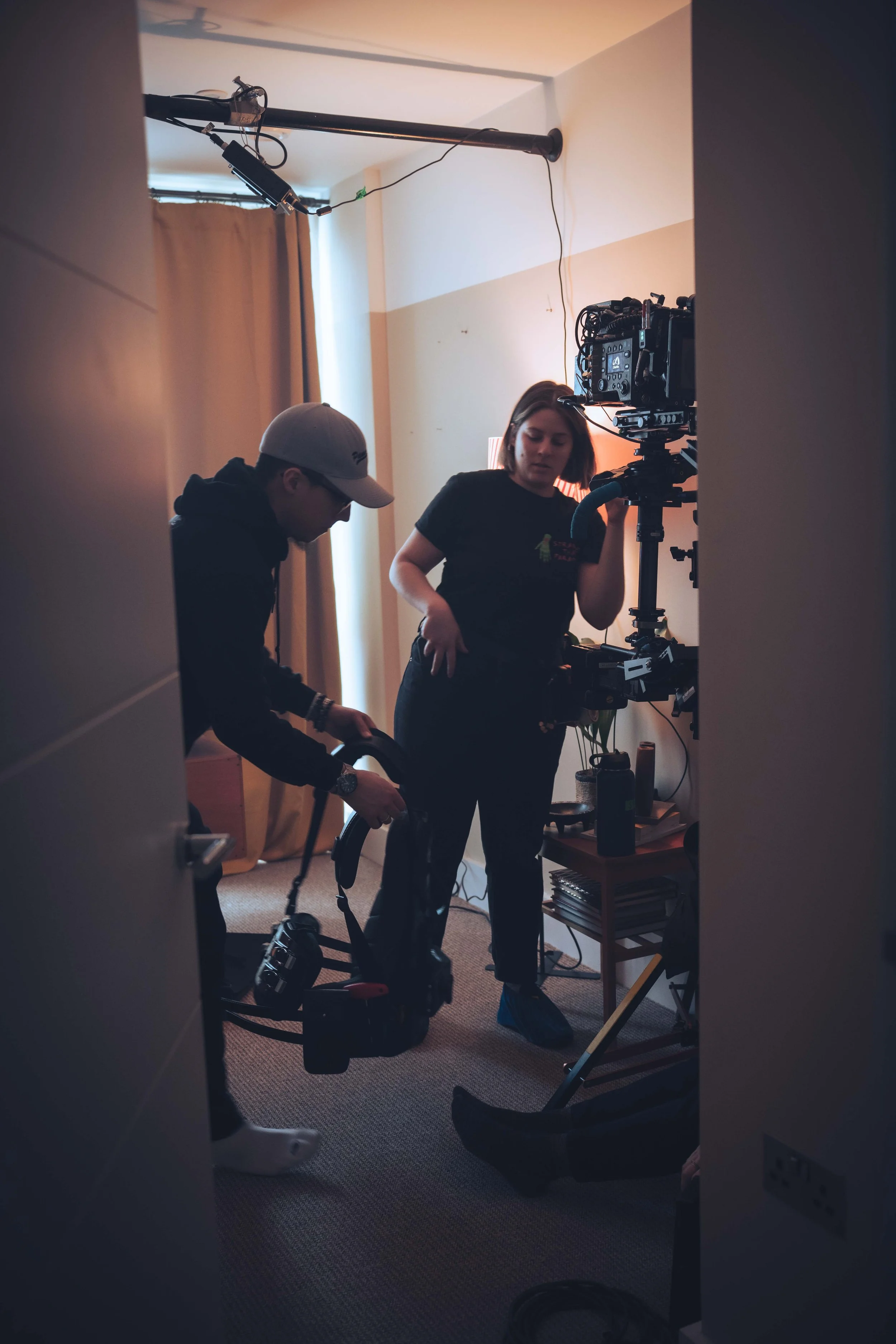 Two people are working with filming equipment in a room. One person is a woman adjusting a camera on a tripod, while the other is a man holding a vacuum cleaner. The room has a window with curtains, and there is a small table with books and a speaker