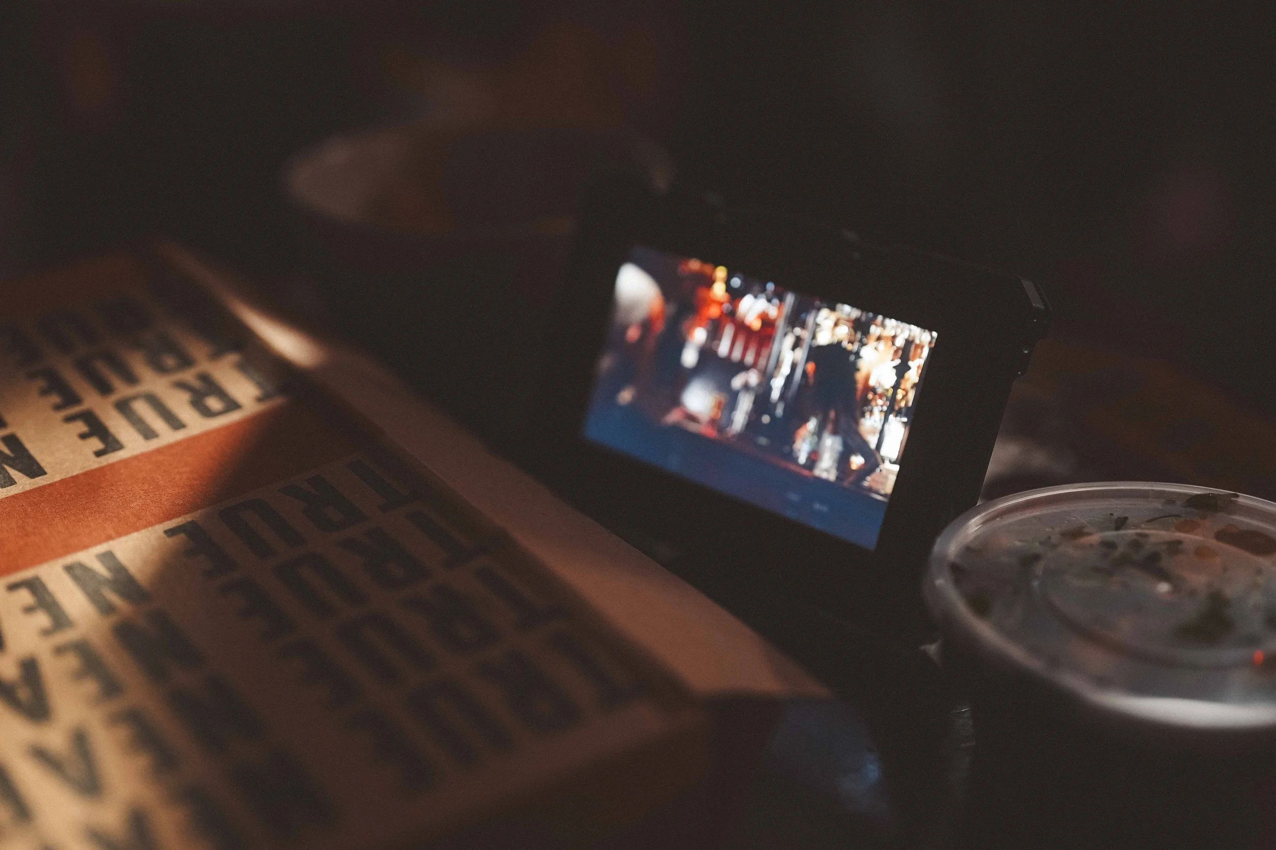 A smartphone on a table capturing a picture, with a pack of cigarettes and a plastic container nearby in a dimly lit environment.