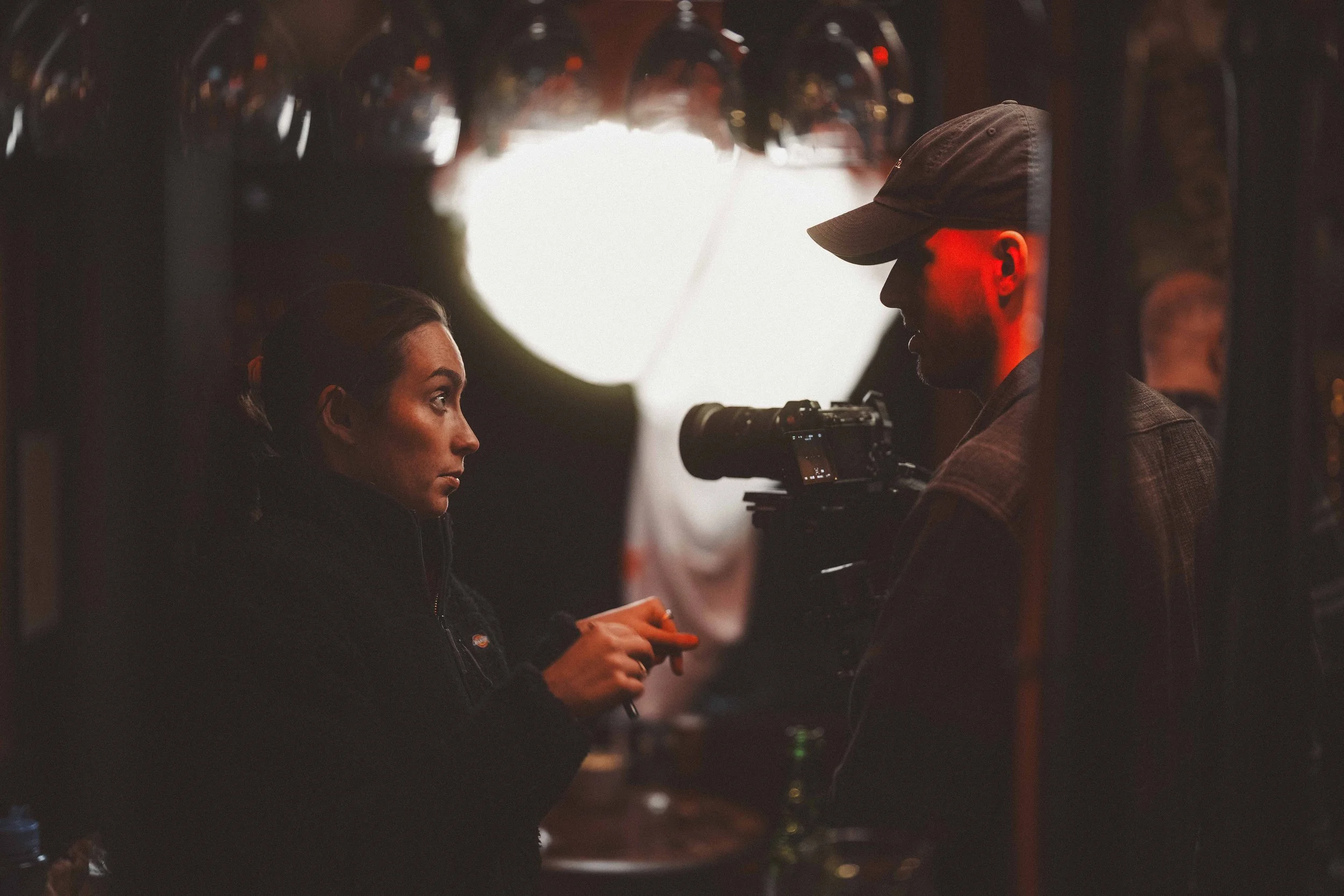 A woman with brown hair and a man with a beard and wearing a cap are looking at each other in a dimly lit room, with a camera on a tripod between them, possibly on a film set.