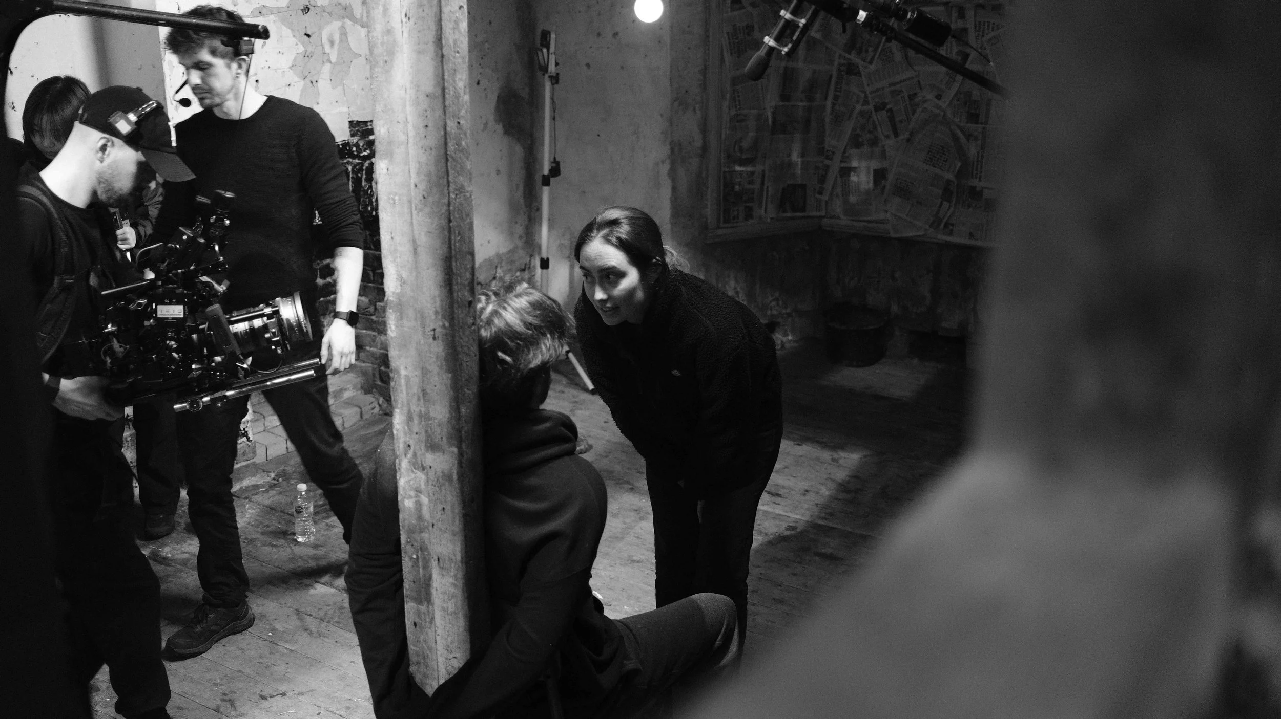 Black and white photo of a film crew on a set. A woman is leaning down towards a seated person, likely giving directions. Several crew members are operating camera equipment, wearing headphones and focused on the scene. The setting is an industrial-s
