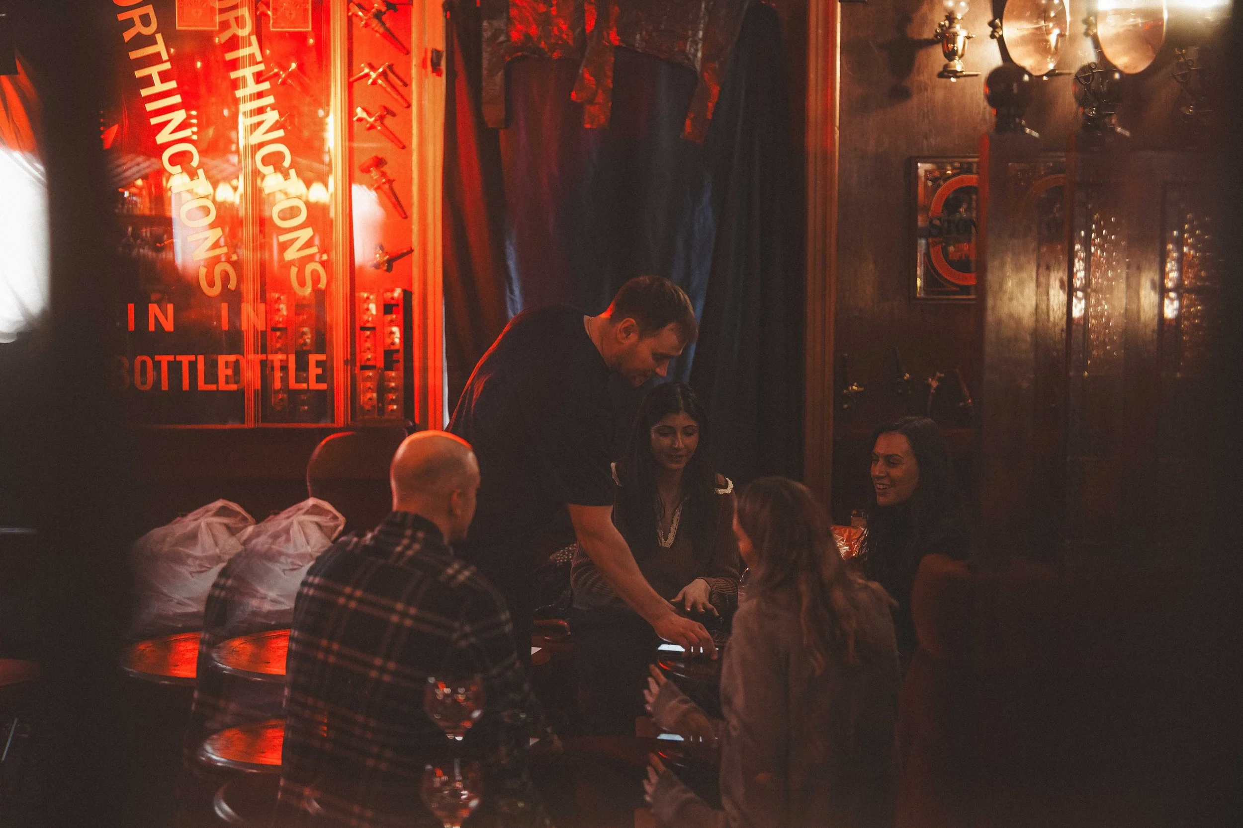 A dimly lit bar or pub with a warm red glow. Several people are gathered, some sitting and some standing, engaged in conversation. The decor includes a display case with liquor bottles and a sign that reads 'THE ONES IN BOTTLE' in red letters. There 
