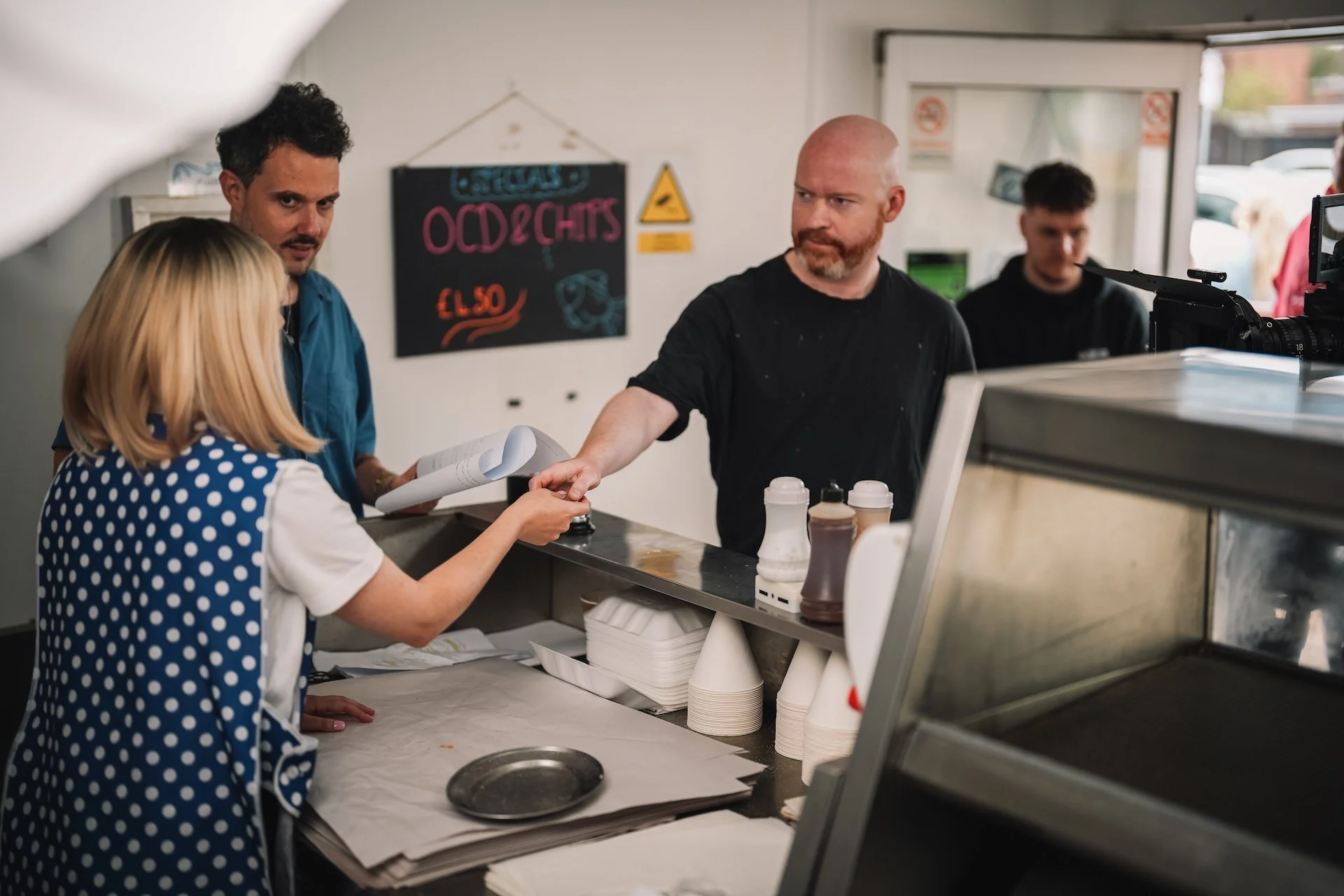 A woman and two men at a counter inside a casual restaurant or cafe, with one man handing her a paper, and a blackboard sign behind them displaying menu items. The woman wears a polka-dot apron, and the man with a beard has a bald head. Another young