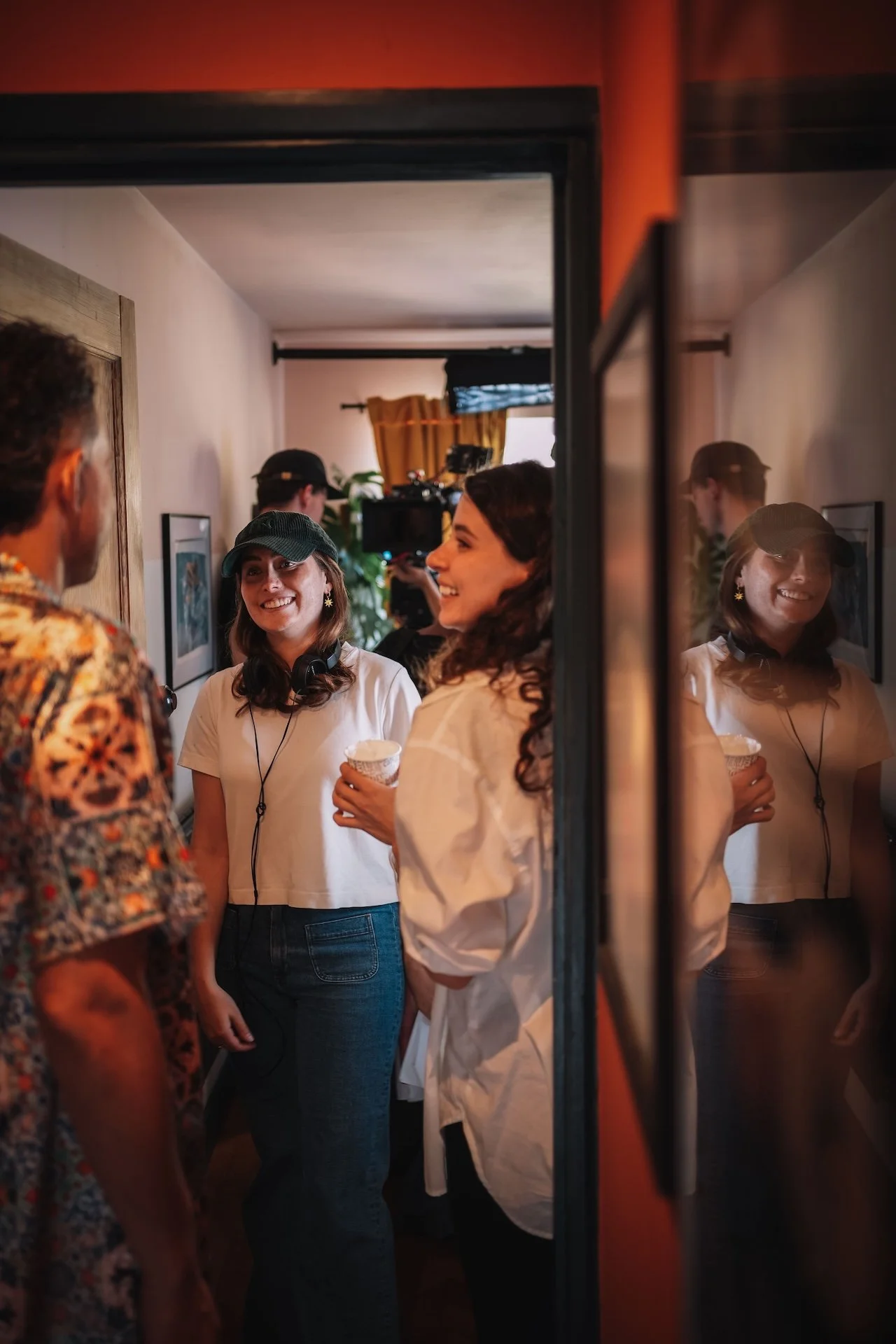 Four women and a man are gathered in a cozy indoor setting, engaging in conversation. The women are smiling, with two of them wearing baseball caps. One woman is holding a cup. A camera crew is visible in the background. The scene is warmly lit with 