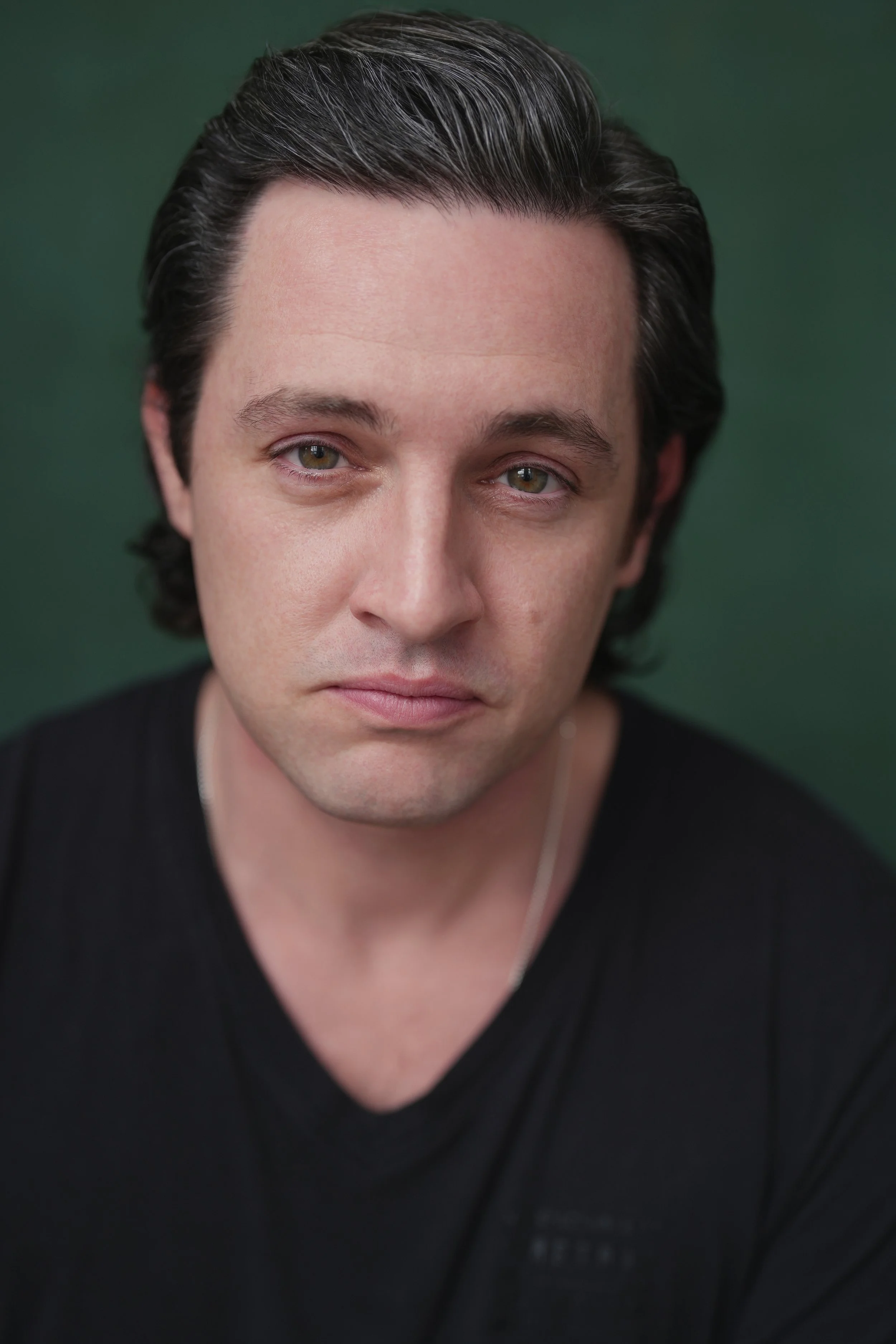 Close-up portrait of a man with dark, slicked-back hair, wearing a black shirt and a thin silver necklace, against a green background.