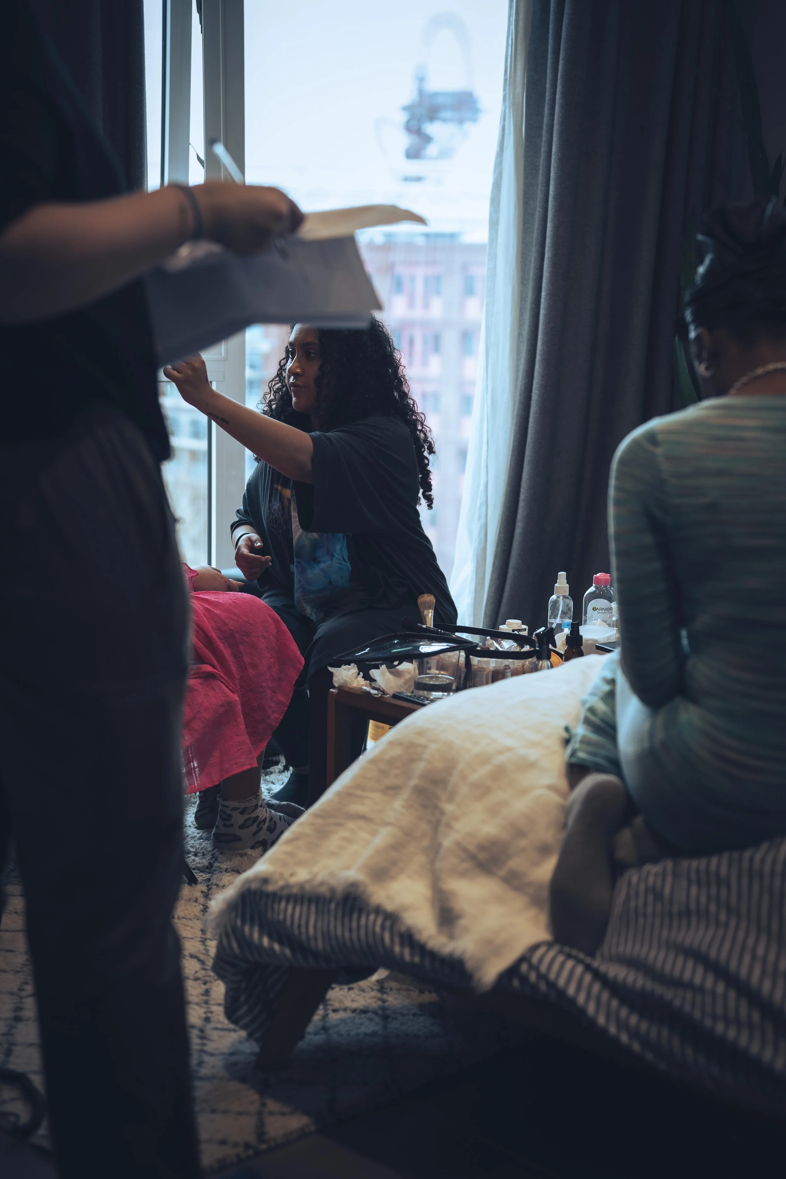 A woman with curly hair and casual clothing is sitting by a window, surrounded by various personal care items and supplies, as another person holds a box or container in the foreground.