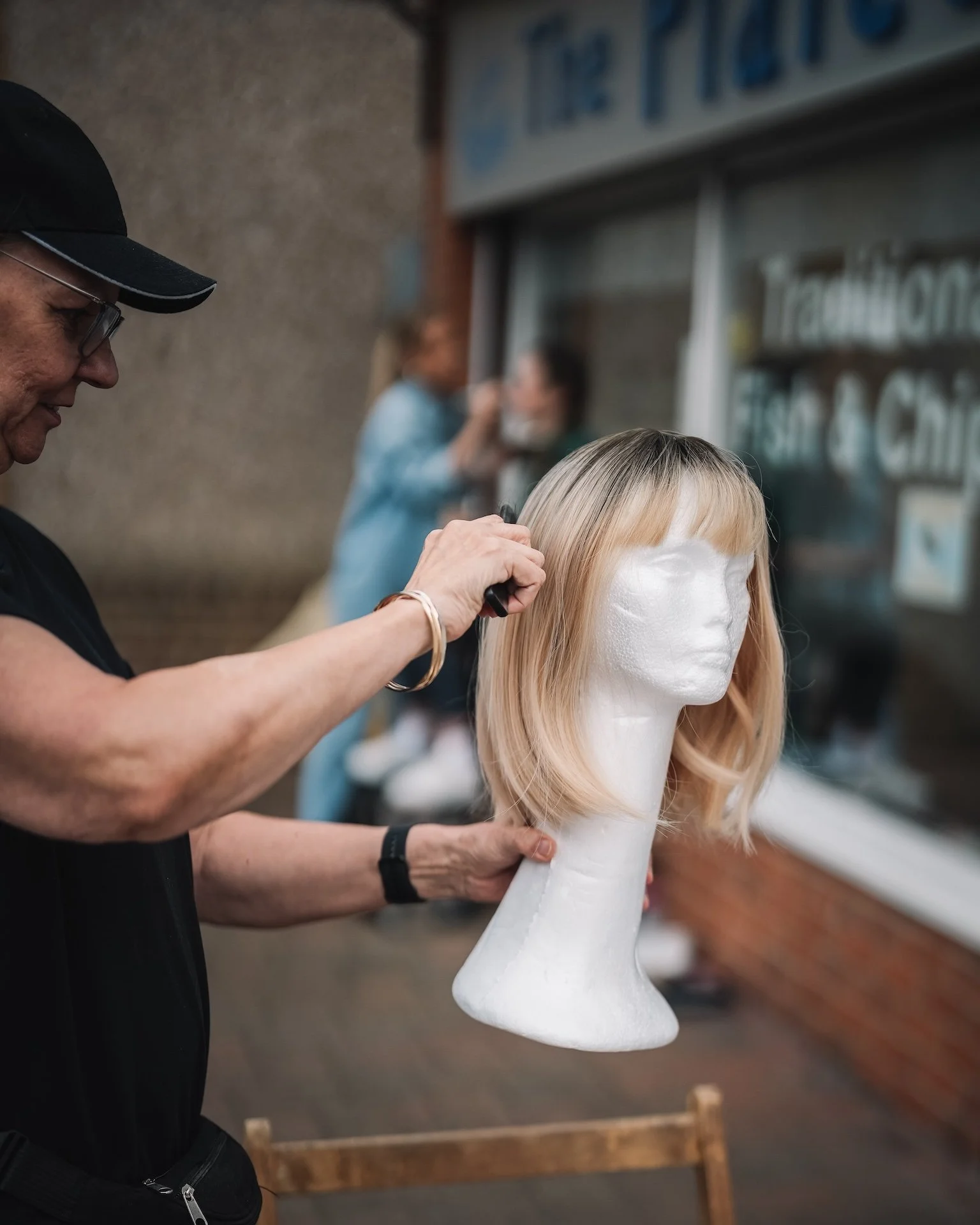 A person is styling a blonde mannequin head with a wig in a salon or cosmetology school setting. The person is wearing glasses and a black hat, focusing on the wig with a comb or styling tool. In the background, another person appears to be working a