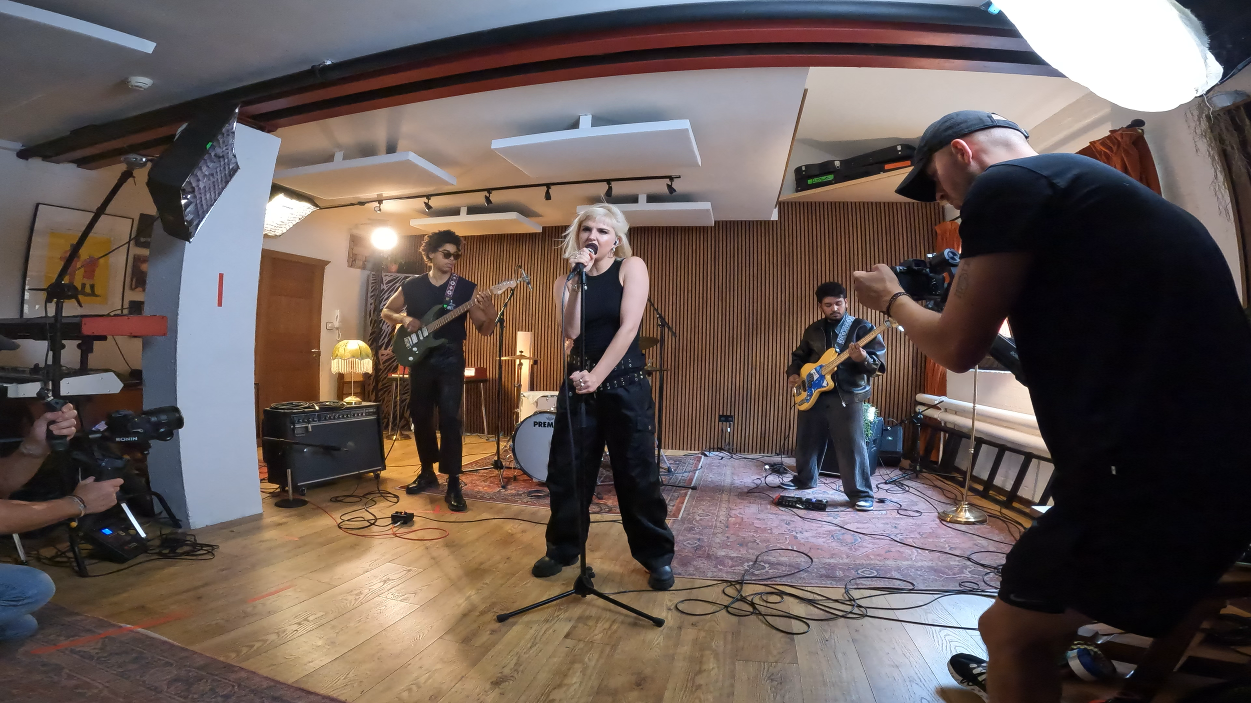 A band performing live in a cozy indoor venue, featuring a female vocalist, two guitarists, and a camera crew capturing the performance.