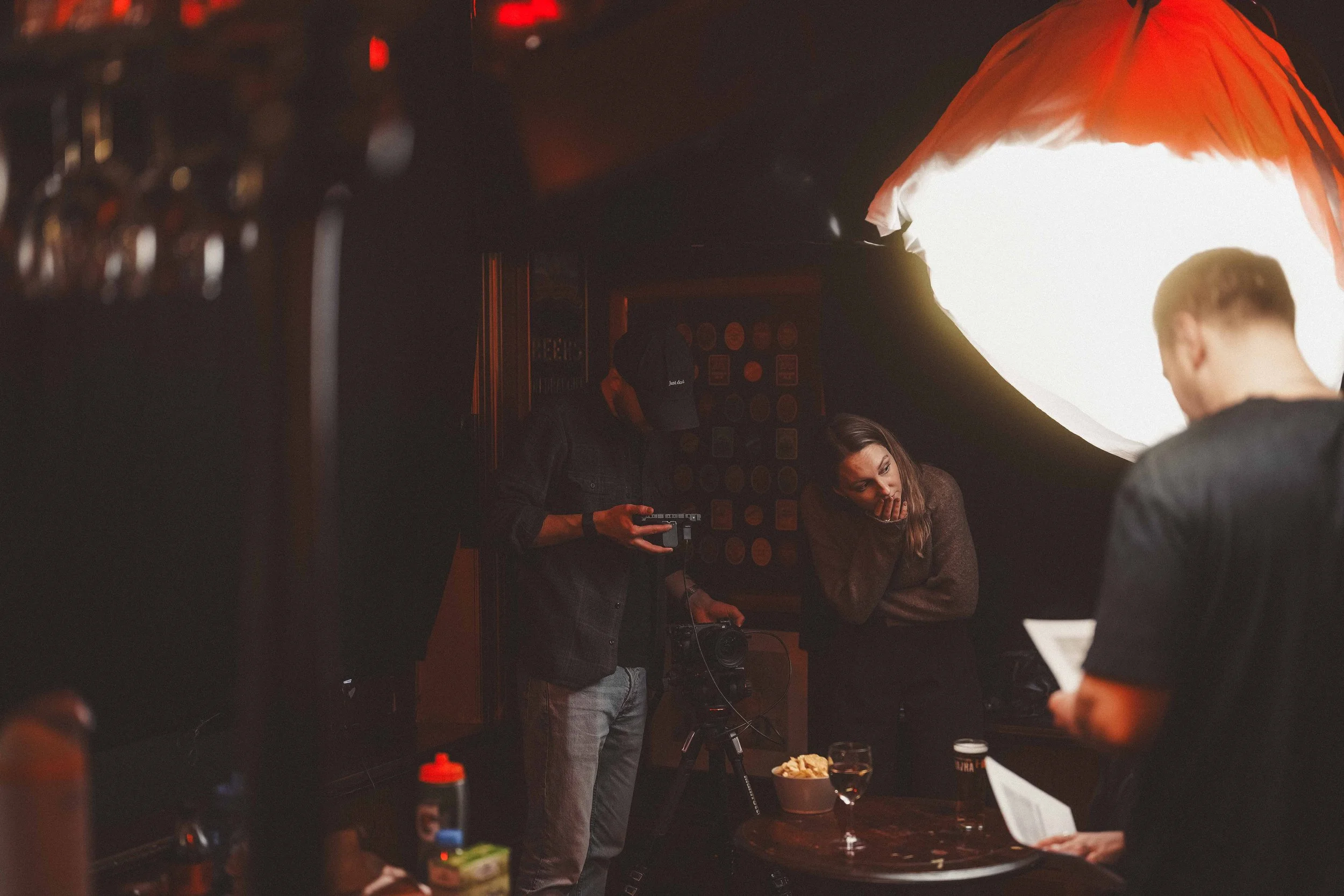 People gathered around a table in a dimly lit setting, possibly a bar or restaurant, with various objects including a bowl of chips, a glass of wine, and some condiments, engaging in conversation.