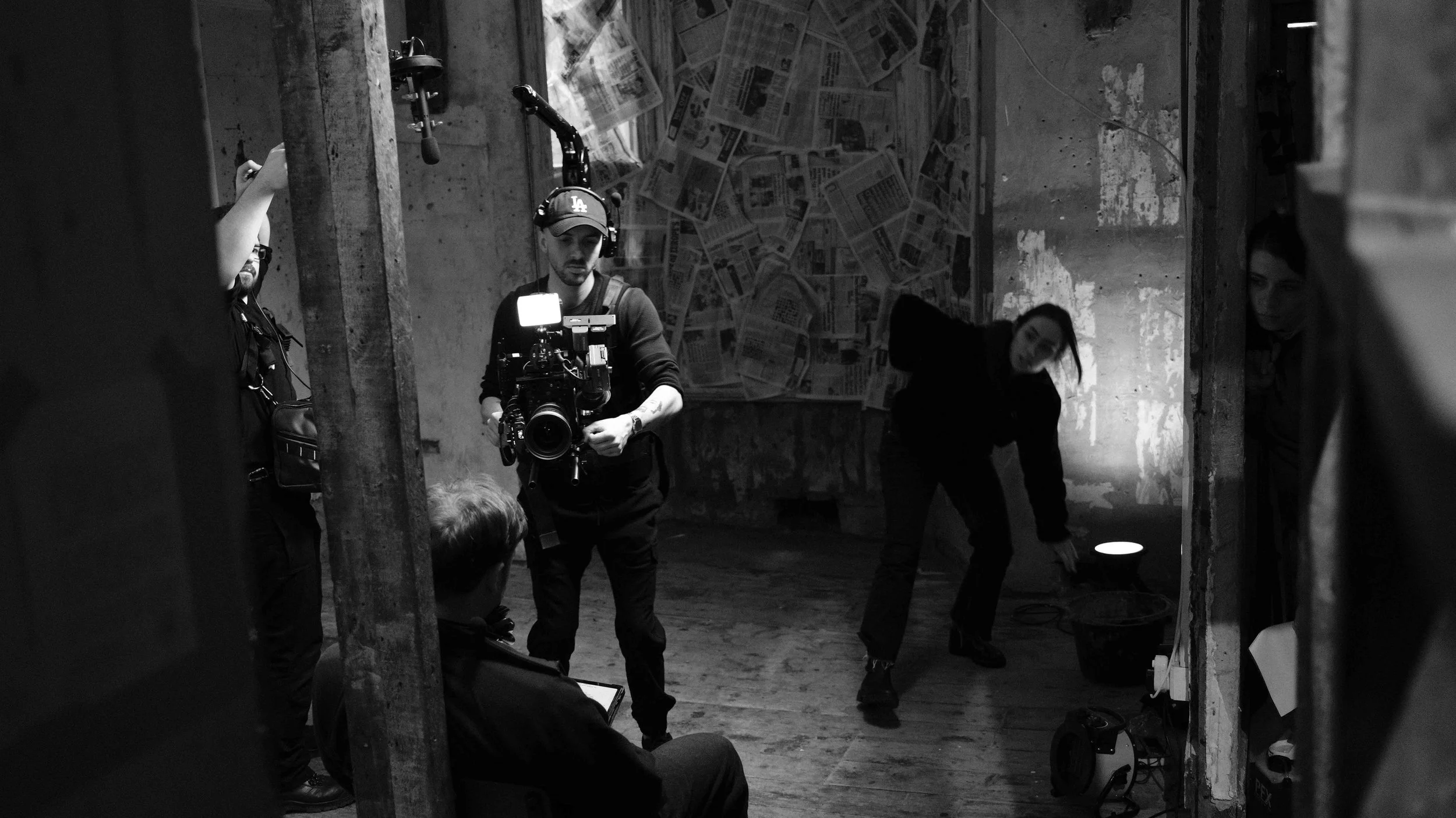 A black-and-white photo of a film crew on set. A man is holding a professional camera, gathered around by others, with one person sitting and two women working on set design or lighting in a rustic, partially unfinished room with newspaper-covered wa