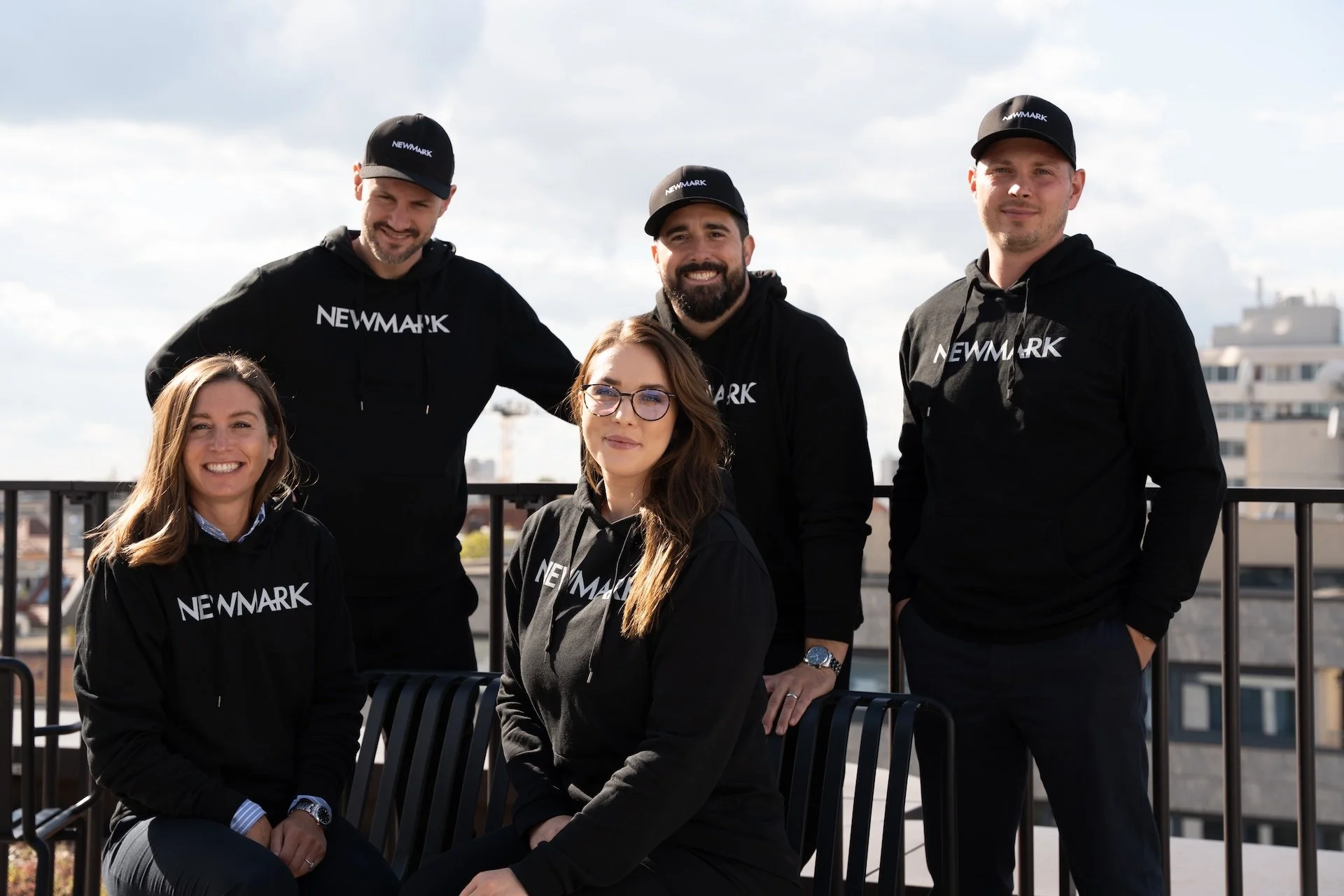 Group of five young professionals wearing black hoodies and caps with 'NEWMARK' logo on a rooftop with city buildings in the background.