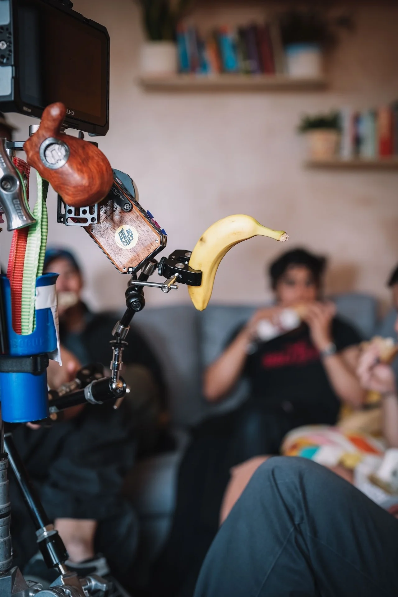 A banana is attached to a tripod-mounted camera or smartphone with a clip, while people in the background are seated on a couch and eating snacks.