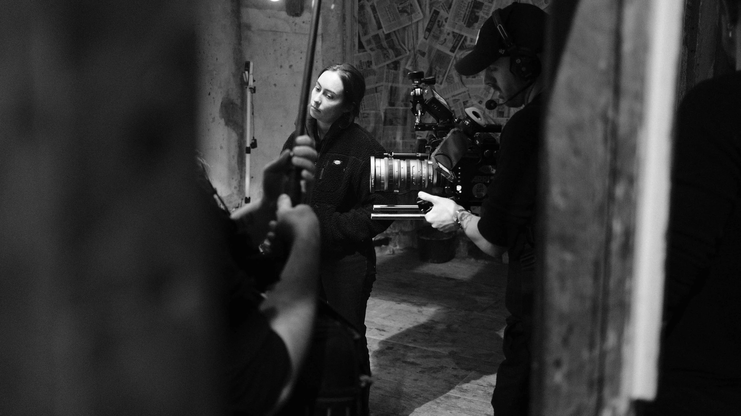A woman in a dark sweater is standing in front of a camera crew in a dimly lit room. The crew, wearing headphones and caps, is setting up equipment, with a large camera pointed at the woman. The background has a wall covered with newspapers.