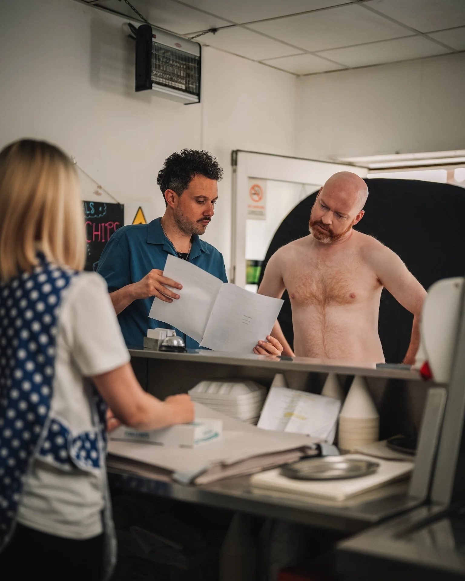 A man with a bald head and beard is shirtless and looking at papers, while another person in a blue shirt is reading from a document. A woman with blonde hair in a polka dot shirt is partially visible, standing at a counter with stacks of papers and 