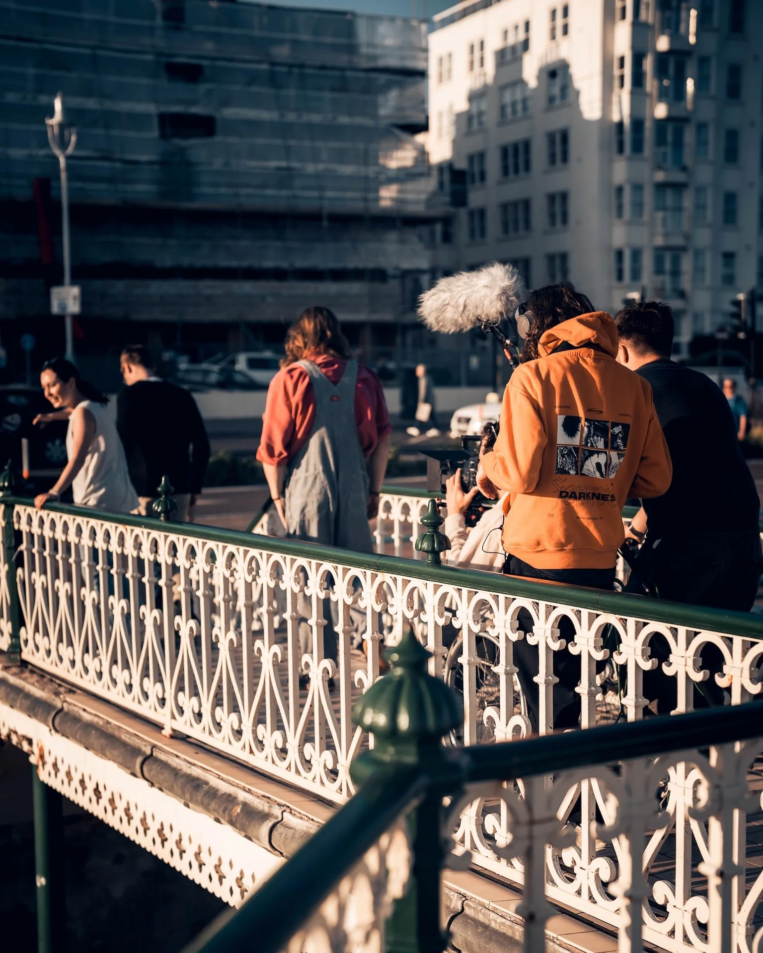 People gathered outdoors near a decorative white fence with urban buildings in the background, some of them engaged in conversation or filming.
