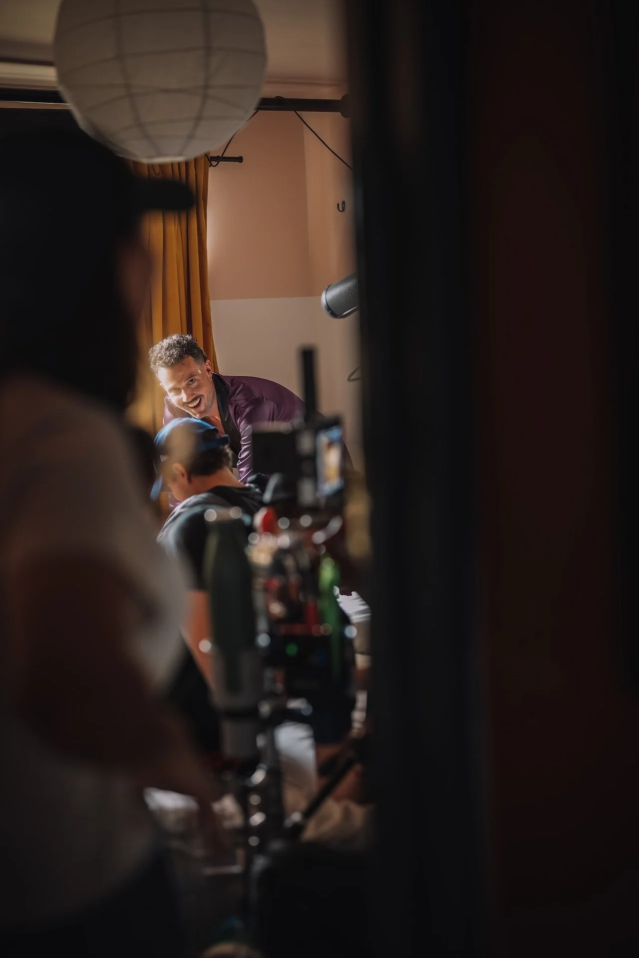 A man with curly hair and a beard is smiling and leaning forward in a room with beige walls and gold curtains. The photo appears to be taken during a film or photo shoot, with equipment and a person operating a camera visible in the foreground.