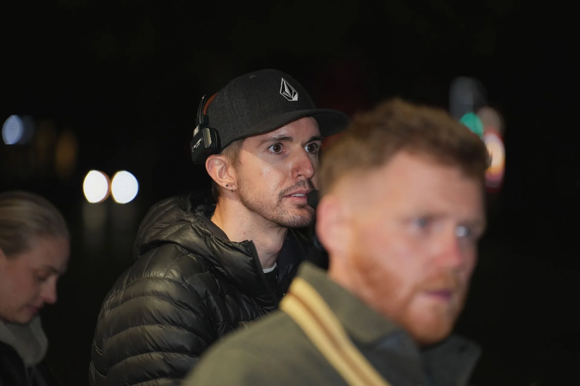A young man with brown eyes and a goatee wearing a black baseball cap with a logo and a black jacket, wearing headphones, seen at night with out-of-focus city lights in the background.