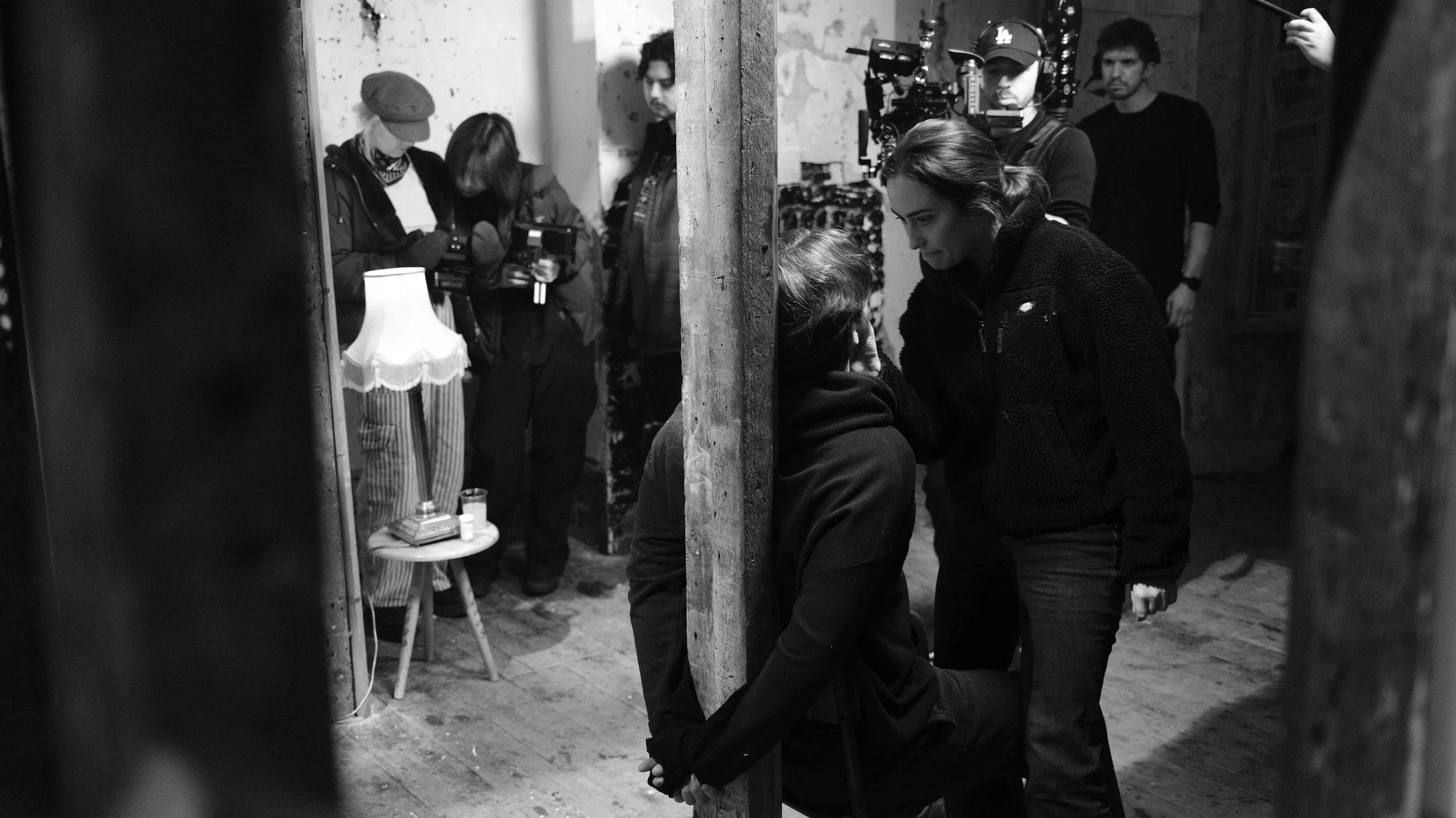 Black and white photo shows a film set scene with two women in foreground, one sitting with hands behind back and head tilted, and the other leaning in close, engaged in conversation. Crew members and a camera operator in the background, some looking