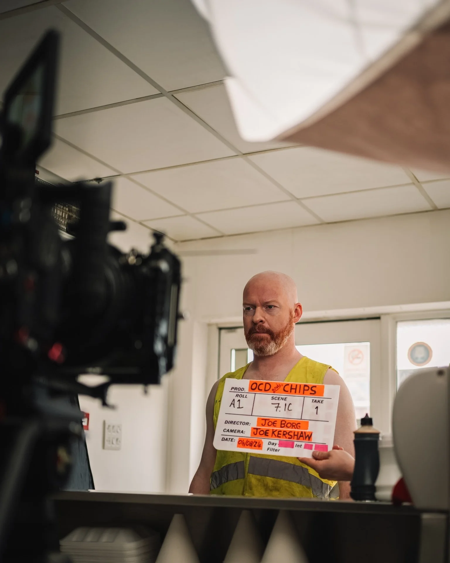 A man with a bald head and red beard holding a clapperboard, standing indoors in front of a window, with a camera and filming equipment in the foreground.