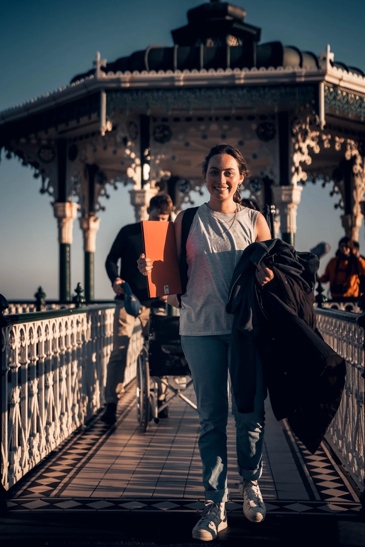 Young woman smiling, holding an orange folder and a backpack, walking on a bridge with a decorative gazebo in the background during sunset.
