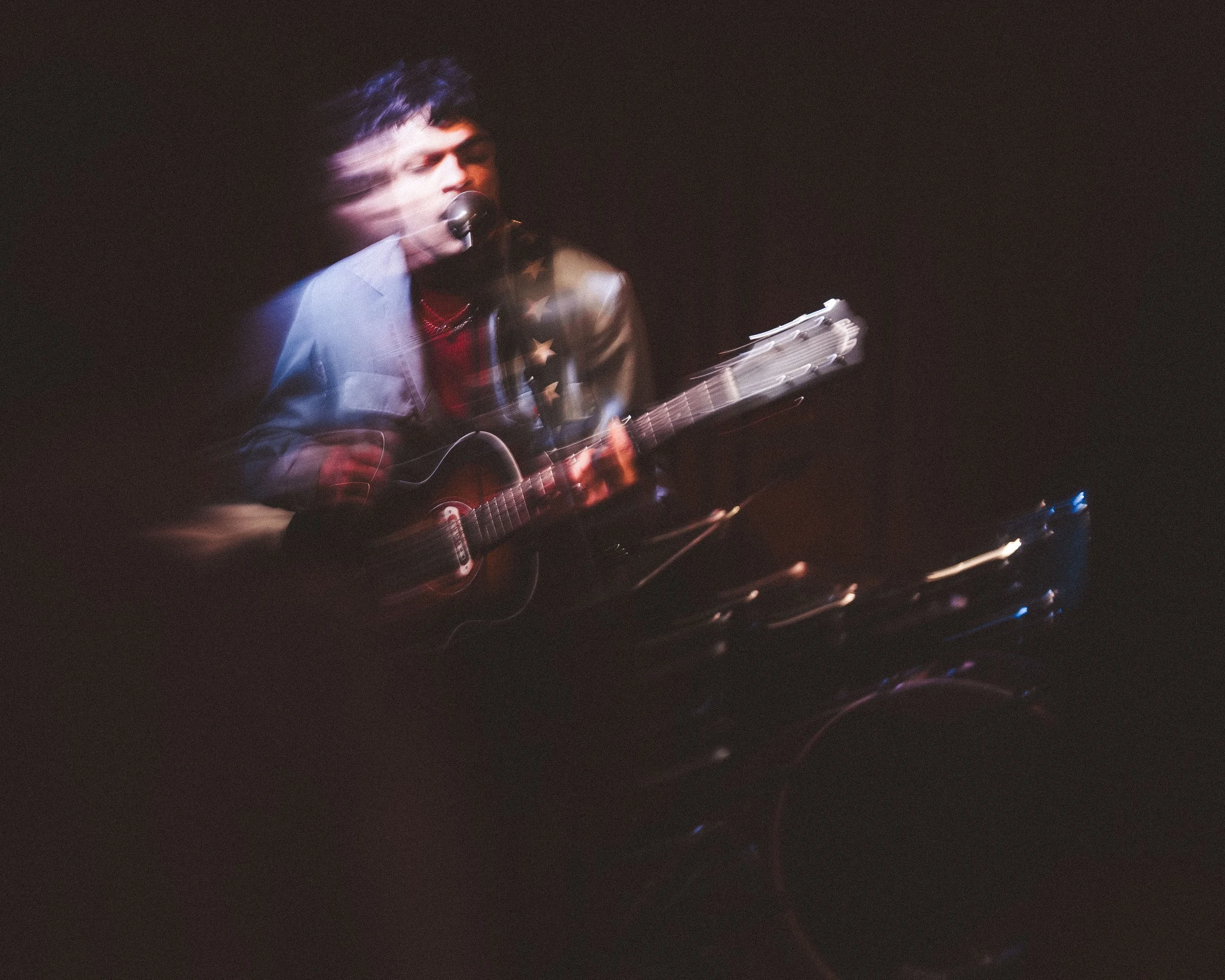 A person playing an acoustic guitar and singing into a microphone, with motion blur effects in a dark setting.