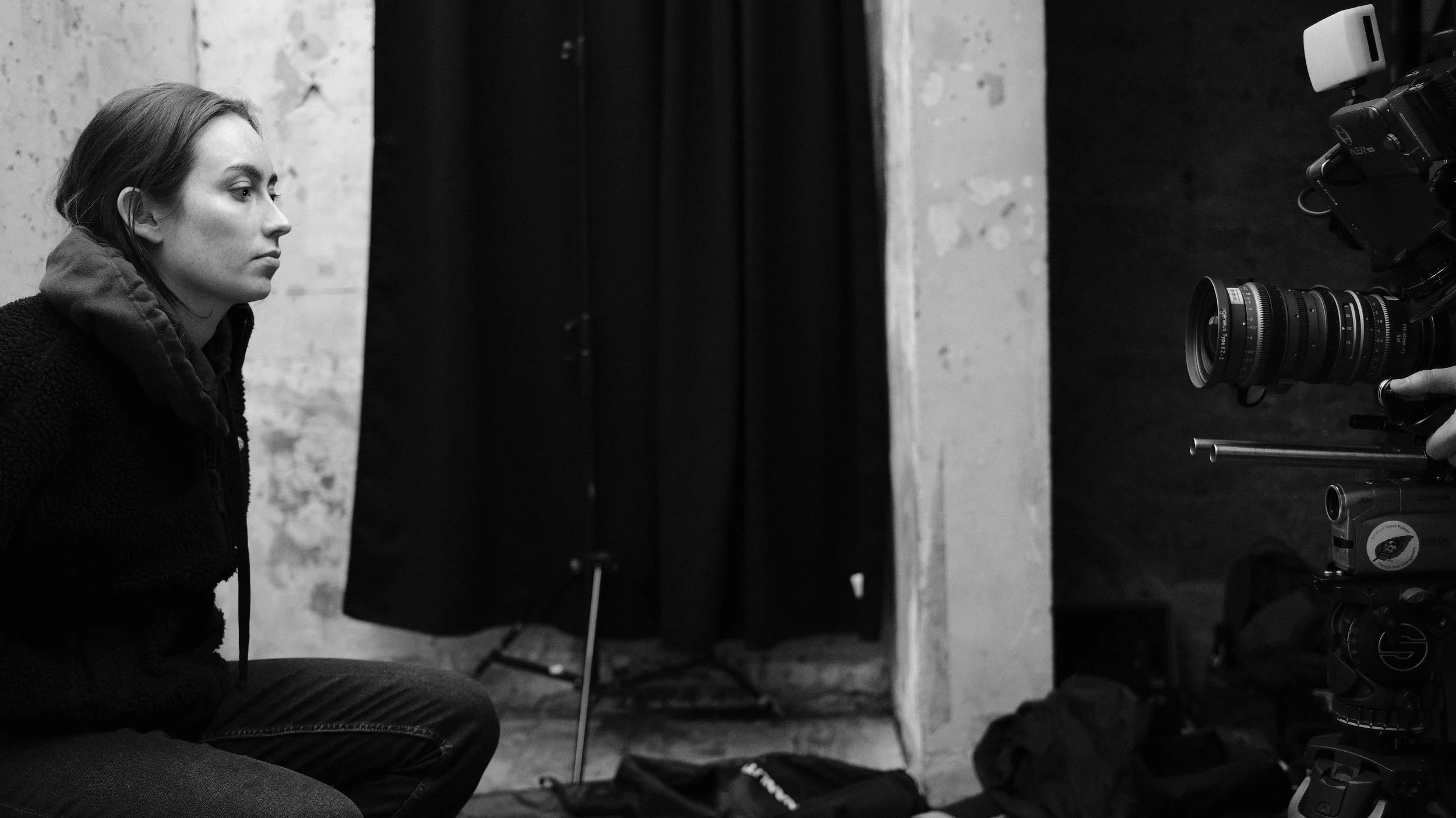 A woman with short hair and a focused expression sits in a photography studio facing a professional camera on a tripod. The studio has a textured wall, a black curtain, and equipment bags on the floor.