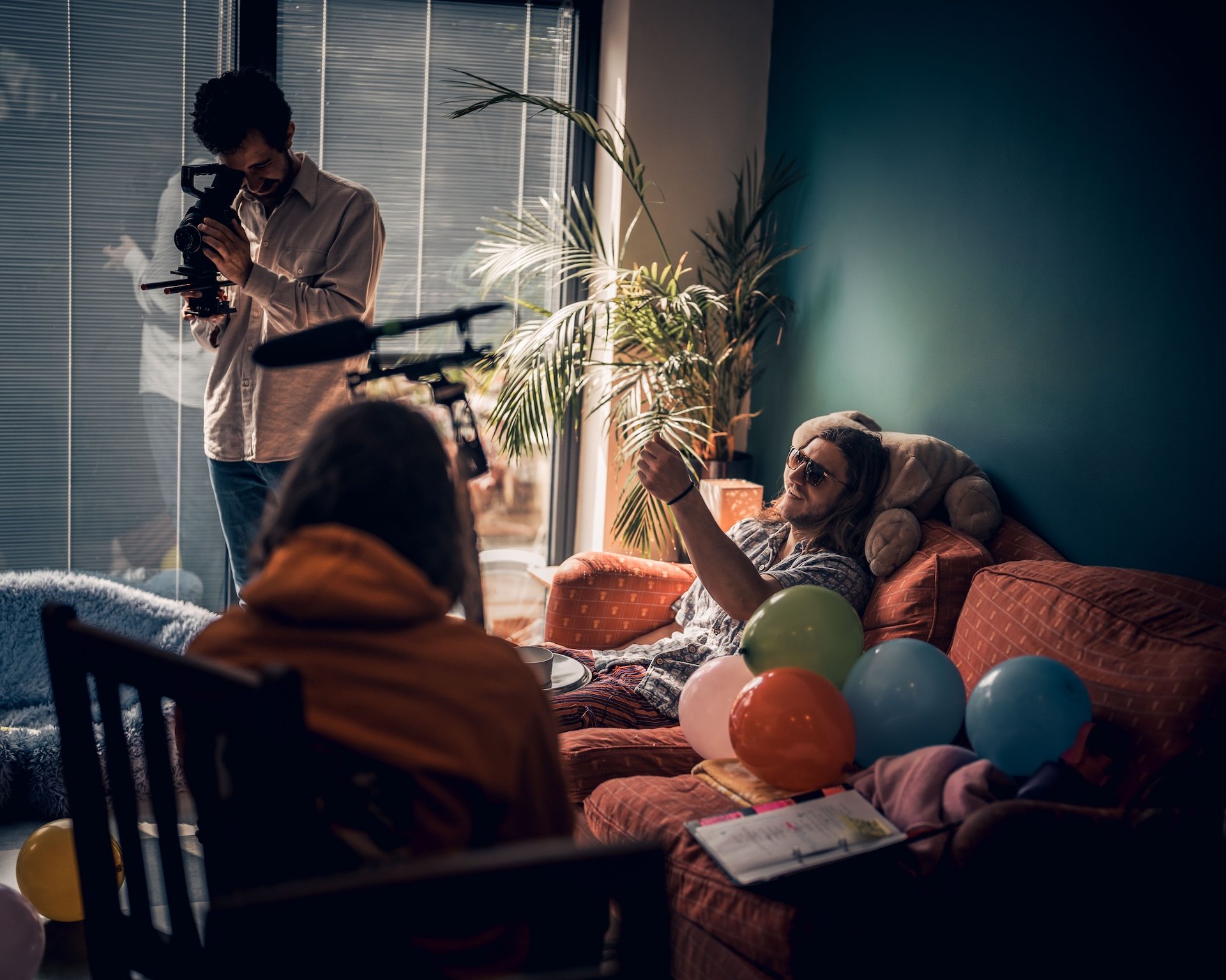 A man sitting on a couch talking to a person standing, a photographer taking pictures, balloons on the couch, and a woman sitting with balloons around, in a room with large windows and plants.